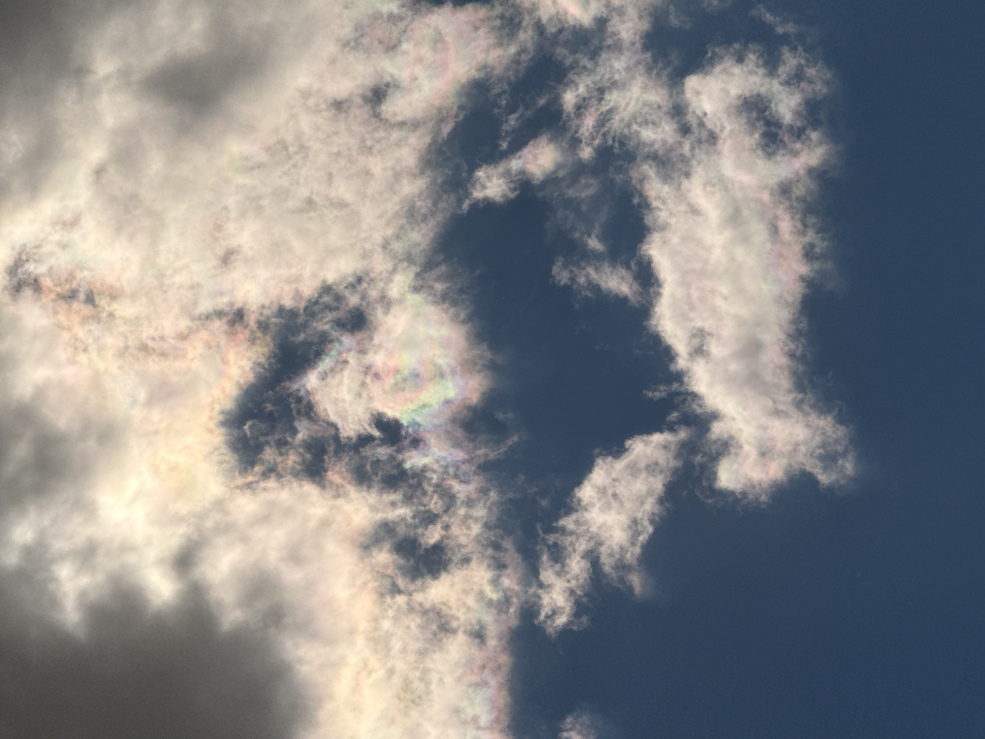 The image captures a sky with clouds displaying iridescence. The cloud formations are predominantly white and gray, with some areas exhibiting a rainbow-like sheen due to diffraction. The background is a deep blue, representing a clear sky. The scene is suggestive of a natural, outdoor setting. The location of Apnagyugh, Armenia, is inferred from the image’s metadata.