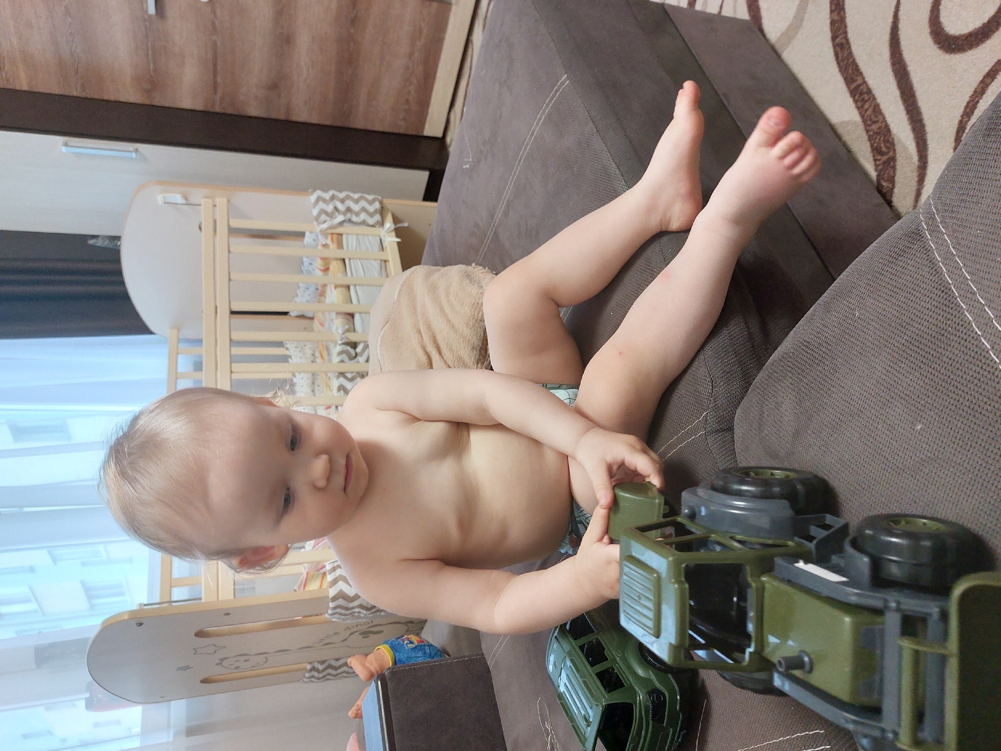 A baby sits on a gray couch, appearing to play with an olive green toy military truck. A baby's crib is visible in the background, along with a wooden piece on the side with drawn shapes on it. The location is likely inside a residence. The city of Chaikovsky, Russia is an inference based on image origin and not directly observable.