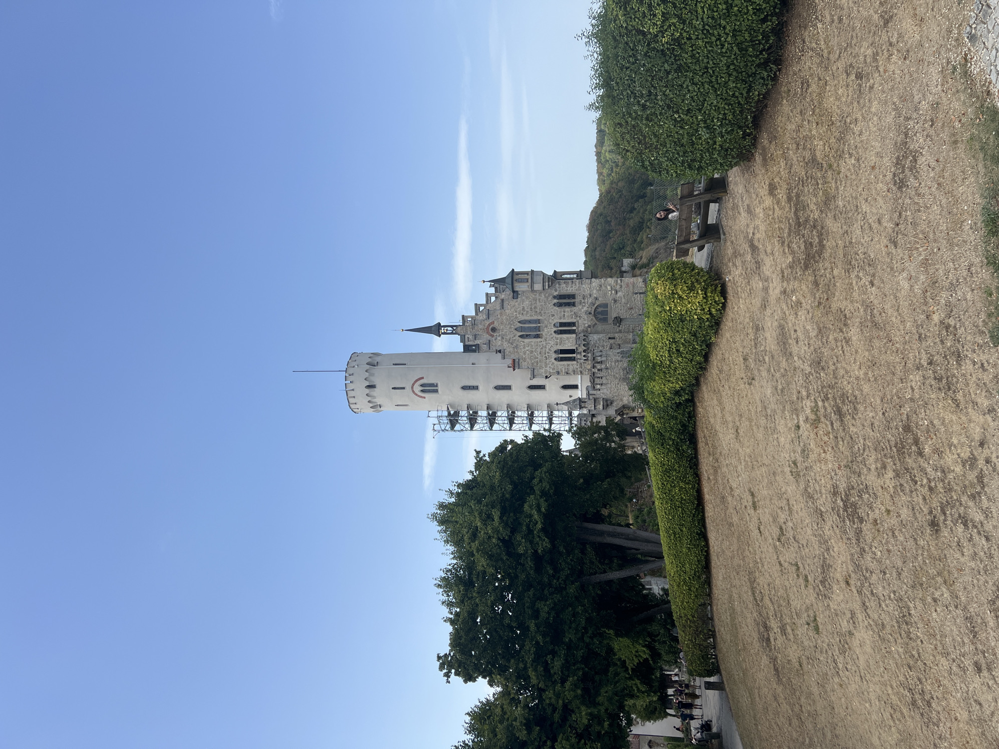 A vertical, wide shot captures Lichtenstein Castle, an architectural marvel set against a clear, pale blue sky. The castle, constructed from stone, features a tall, cylindrical white tower with crenelations and a smaller, spire-topped tower. Scaffolding is visible on the side of the white tower, suggesting ongoing restoration work. Below the main structures, a stone wall with battlements forms the base of the castle.
In the foreground, a grassy slope, dried and somewhat sparse, leads up to a neatly trimmed green hedge. To the left of the castle, a cluster of large, leafy green trees provides shade. Beyond the castle, a lush, tree-covered hillside descends into a valley. A wooden bench sits to the right, where a young woman with dark hair, wearing a light-colored jacket, is seated and looking at her phone. Several people are visible walking on a paved path to the left of the castle, in front of a white building. The overall scene conveys a sense of historical grandeur and natural beauty under a bright, sunny day.