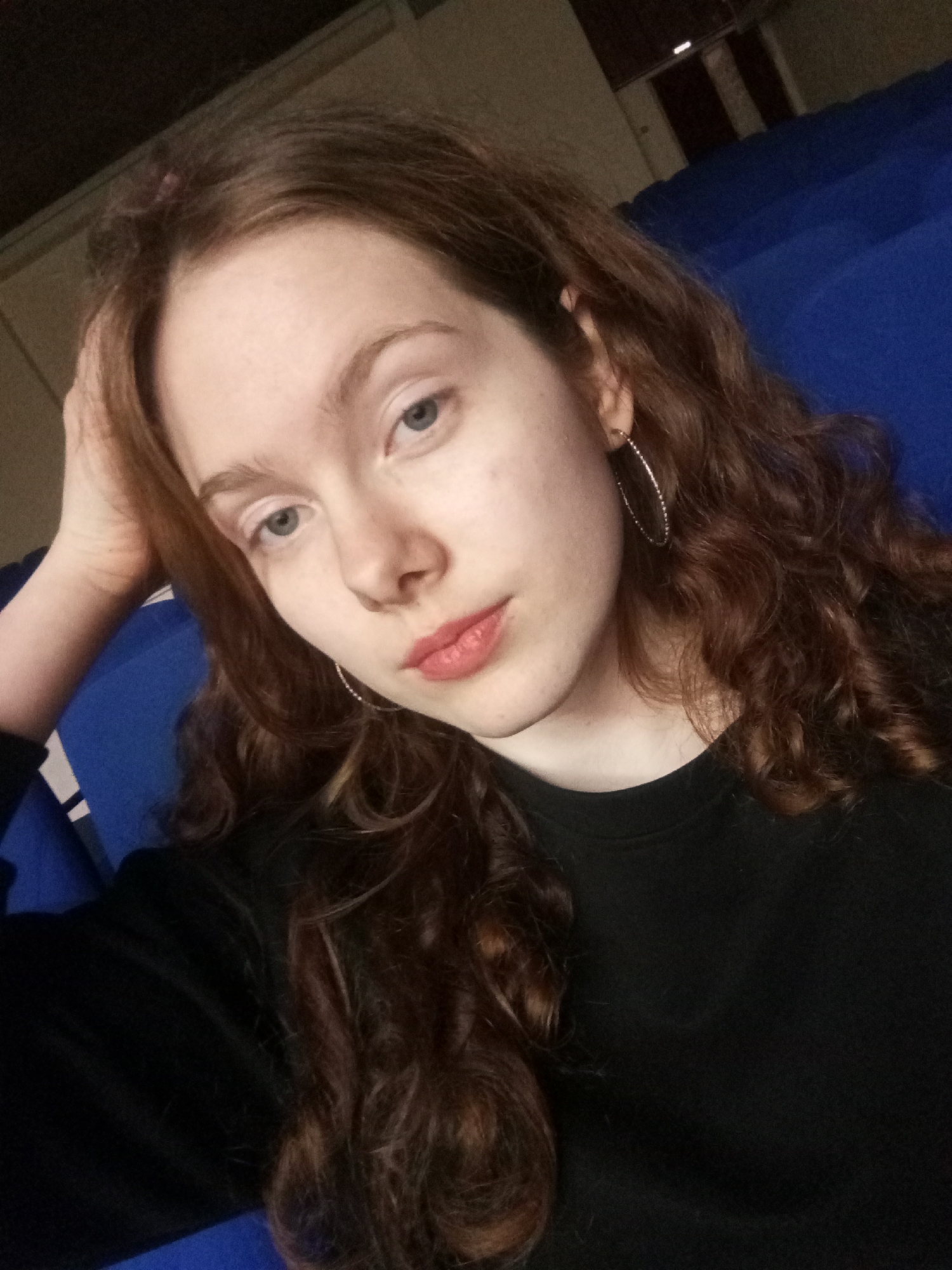 A young woman with long, wavy, auburn hair and fair skin is the subject of a selfie-style photograph. She is wearing a black, long-sleeved shirt and silver hoop earrings. Her face is angled slightly to the left, and her head is resting on her hand. She has light blue eyes and is wearing light pink lipstick. The background features blue, upholstered seating, possibly in an auditorium or theater. A dimly lit corridor is visible in the upper right corner, suggesting an indoor location. Without additional information, determining the exact location in Chaikovsky, Russia is impossible. The image presents a candid and casual portrait in an indoor setting.