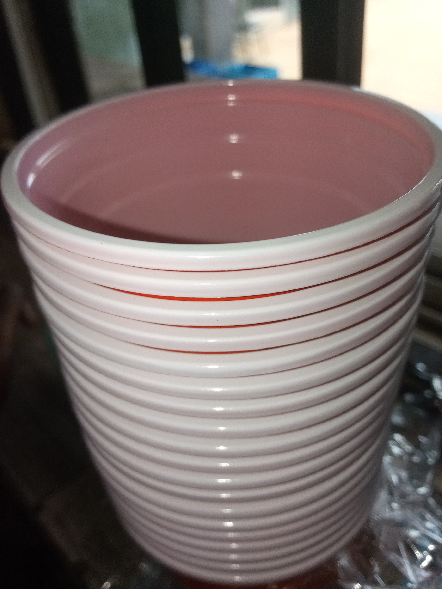 A close-up, vertical, eye-level shot shows the exterior of a pink ceramic pot with a ribbed design and red accents. The pot is cylindrical and wider at the top than the bottom. The interior is a soft pink color, while the exterior is a lighter pink with horizontal grooves. Thin red lines are visible in some of the grooves, creating a striped effect. The background is blurred, showing dark vertical lines and lighter areas, suggesting an indoor environment with some natural light. The scene appears to be a still life of the decorative pot.