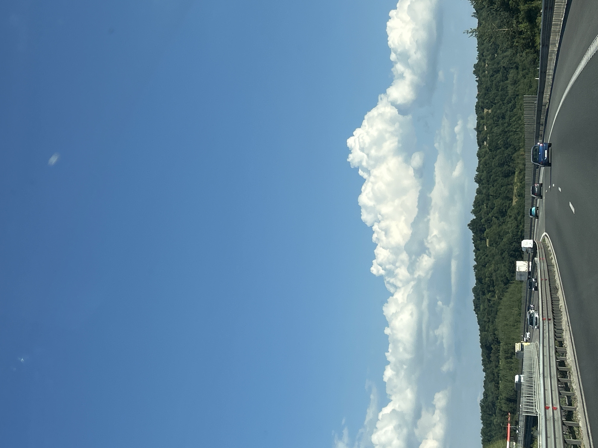 The image depicts a highway scene with cars and trucks traveling under a bright blue sky with scattered white clouds. The road curves to the right, bordered by metal guardrails. Beyond the highway, a dense, green forest covers rolling hills. The context of Baliovtsi, Bulgaria, suggests this is a view of a major roadway likely connecting or passing through the region. The image is captured from a moving vehicle, as indicated by the perspective and the motion blur present in some elements. There are no visible people in the image. The overall scene is characteristic of a sunny day on a highway in a rural or semi-rural area.