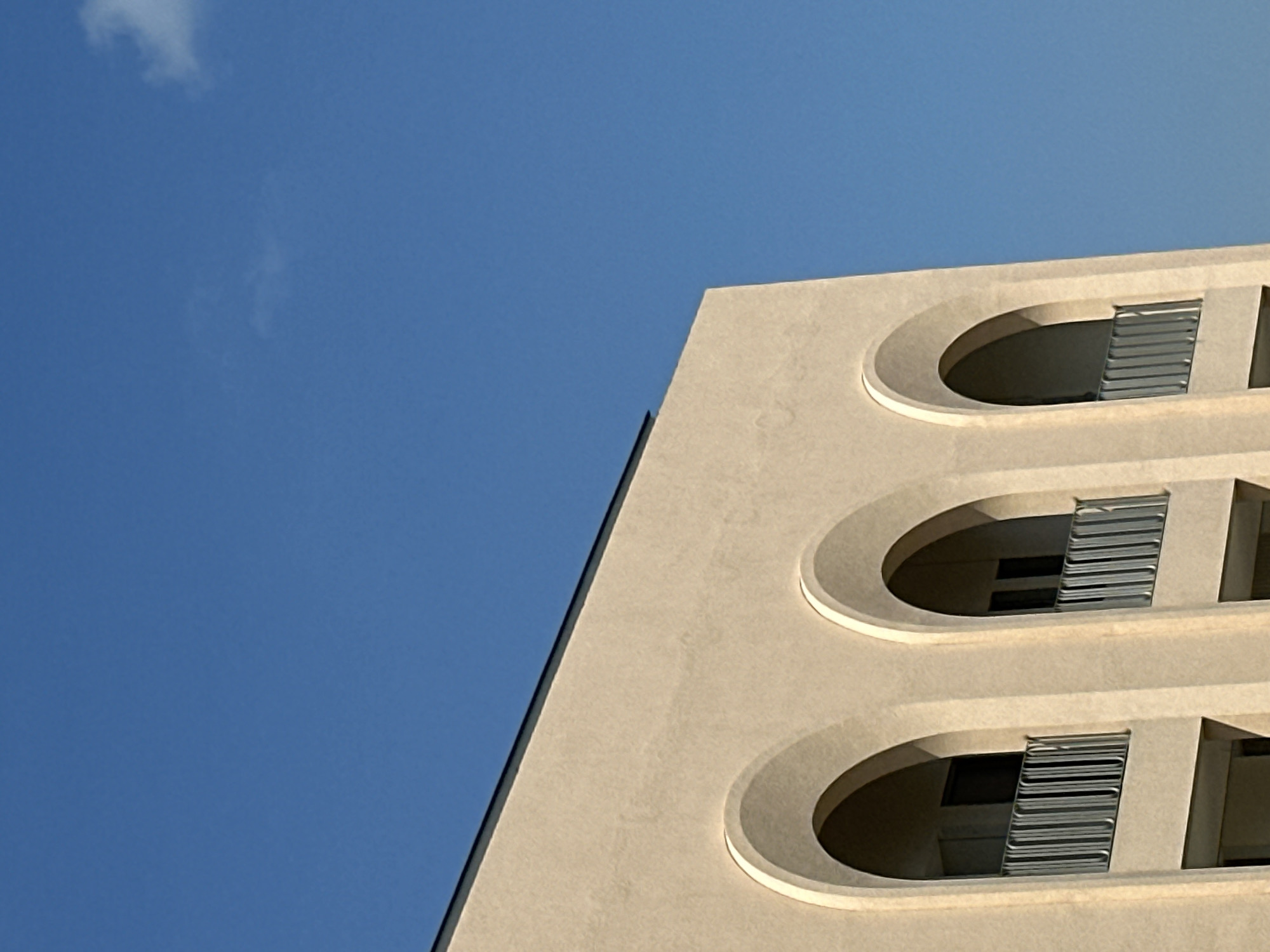 The image captures a close-up, low-angle view of a modern building with a light beige facade against a clear blue sky. Three arched windows with balconies are visible, each featuring a metallic railing. The building appears to be a multi-story structure, with darker recessed areas behind the balconies suggesting interior spaces. The architectural style is clean and contemporary. The sky is a vibrant blue with a faint wisp of a cloud visible in the upper right portion of the frame. The lighting suggests it is daytime, likely late morning or early afternoon, given the brightness and the angle of the light. The image does not contain any people or animals. The setting appears to be an urban environment, specifically in the city of North Kuta, Indonesia, given the provided context.