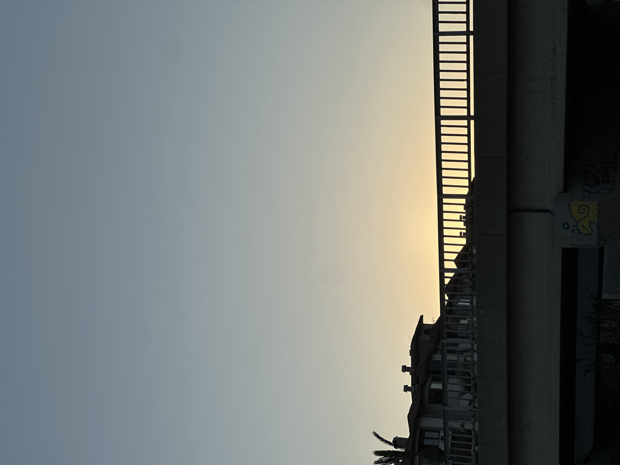 The image depicts a sunset view from under a bridge in Konyaaltı, Turkey. The sky is a gradient of soft blue and yellow, indicating the sun is low on the horizon. A silhouetted bridge railing with vertical bars runs across the middle of the frame, with a multi-story building and a palm tree visible behind it on the left. The lower part of the image is dominated by the dark underside of the bridge, showing some graffiti on a concrete pillar.