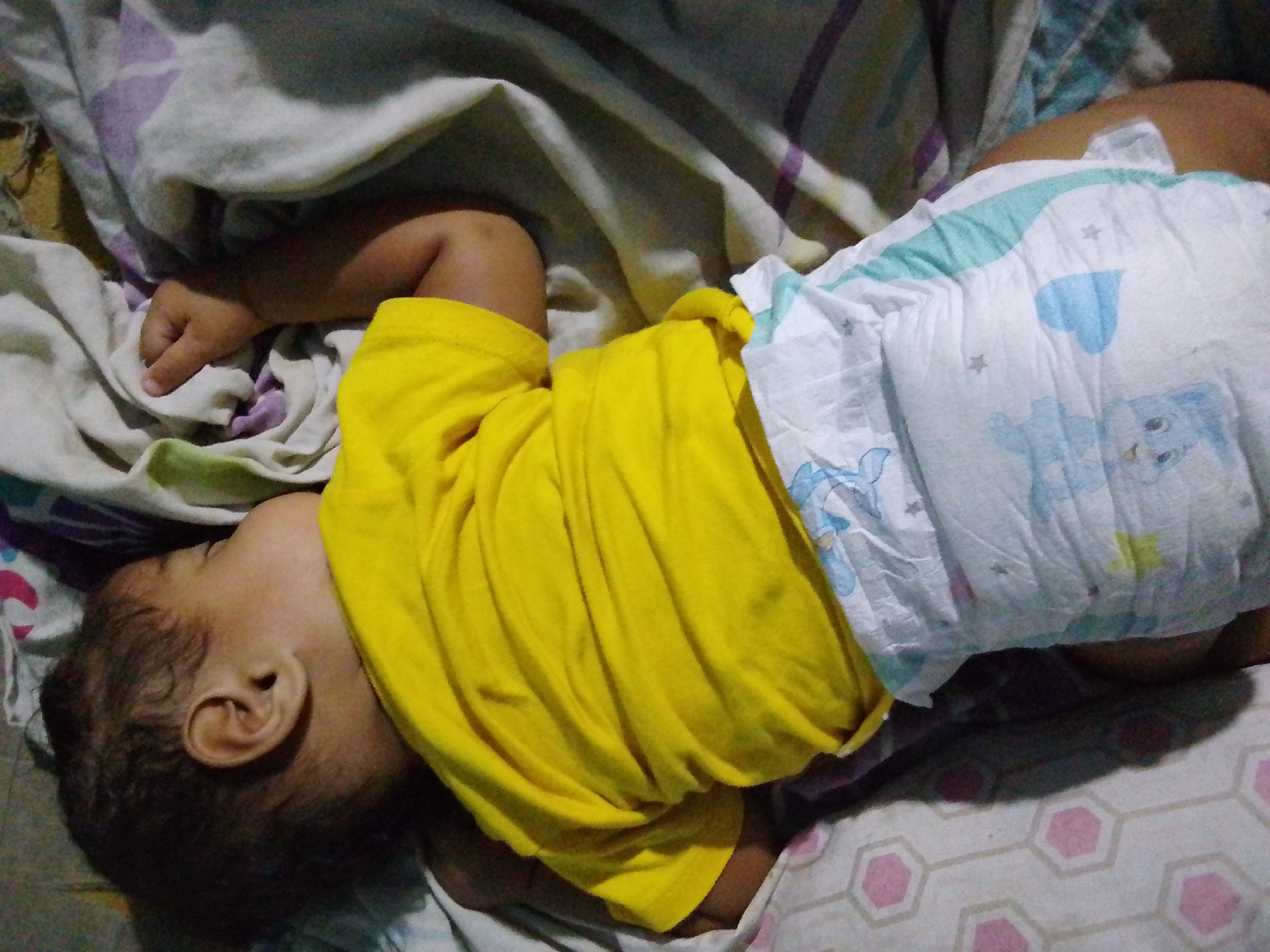 A baby is captured in a close-up, high-angle shot, lying on its left side and appearing to be asleep or resting. The infant is wearing a vibrant yellow short-sleeved t-shirt and a white disposable diaper. The diaper features light blue accents, including patterns of small stars and what appears to be a cartoon-like animal character, possibly a bear. It looks somewhat full or crinkled, suggesting it might be soiled.
The baby's head is turned away from the camera, revealing the back of its head with dark, curly hair and a visible ear. Its right arm is bent, with a small hand gently resting on a crumpled white cloth or blanket. The baby is lying on patterned bedding; one section is a greyish-white fabric with subtle purple lines or abstract patterns, while another section, partly visible beneath the baby and to the bottom right, has a white background with repeating pink hexagonal shapes.
The overall scene suggests a calm, indoor setting, likely a bedroom or nursery. The lighting is soft, not indicating a specific time of day beyond being an indoor environment. No specific location cues or text are visible in the image. The baby's relaxed posture conveys a sense of peaceful slumber.
