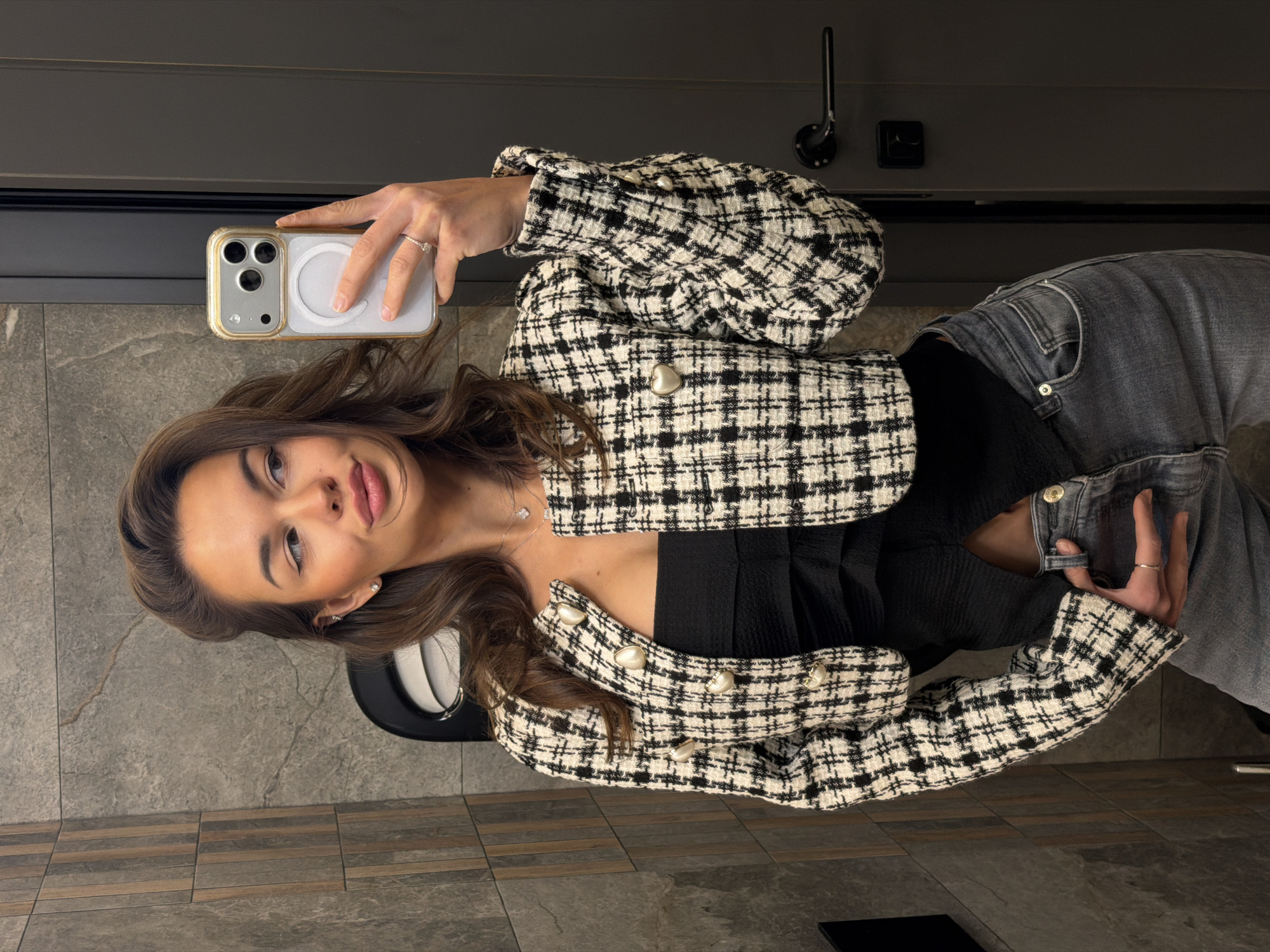 A young woman is taking a selfie in what appears to be a bathroom or restroom. She is holding a smartphone in her left hand, showcasing the phone's camera and case. The woman is wearing a black and white tweed jacket with pearl-like buttons, a black top underneath, and gray jeans. She has brown hair styled with waves and is wearing jewelry, including a ring and earrings.
The background consists of tiled walls and flooring, suggesting a modern restroom setting. Above her is a dark-colored wall or door with a metallic hook and a small black square, possibly a latch or a sensor. In the bottom corner, there is a small black rectangular object. The overall lighting is even and seems to be coming from overhead sources.
It is impossible to confirm that the location is in Krasnoyarsk, Russia, without additional information or context.