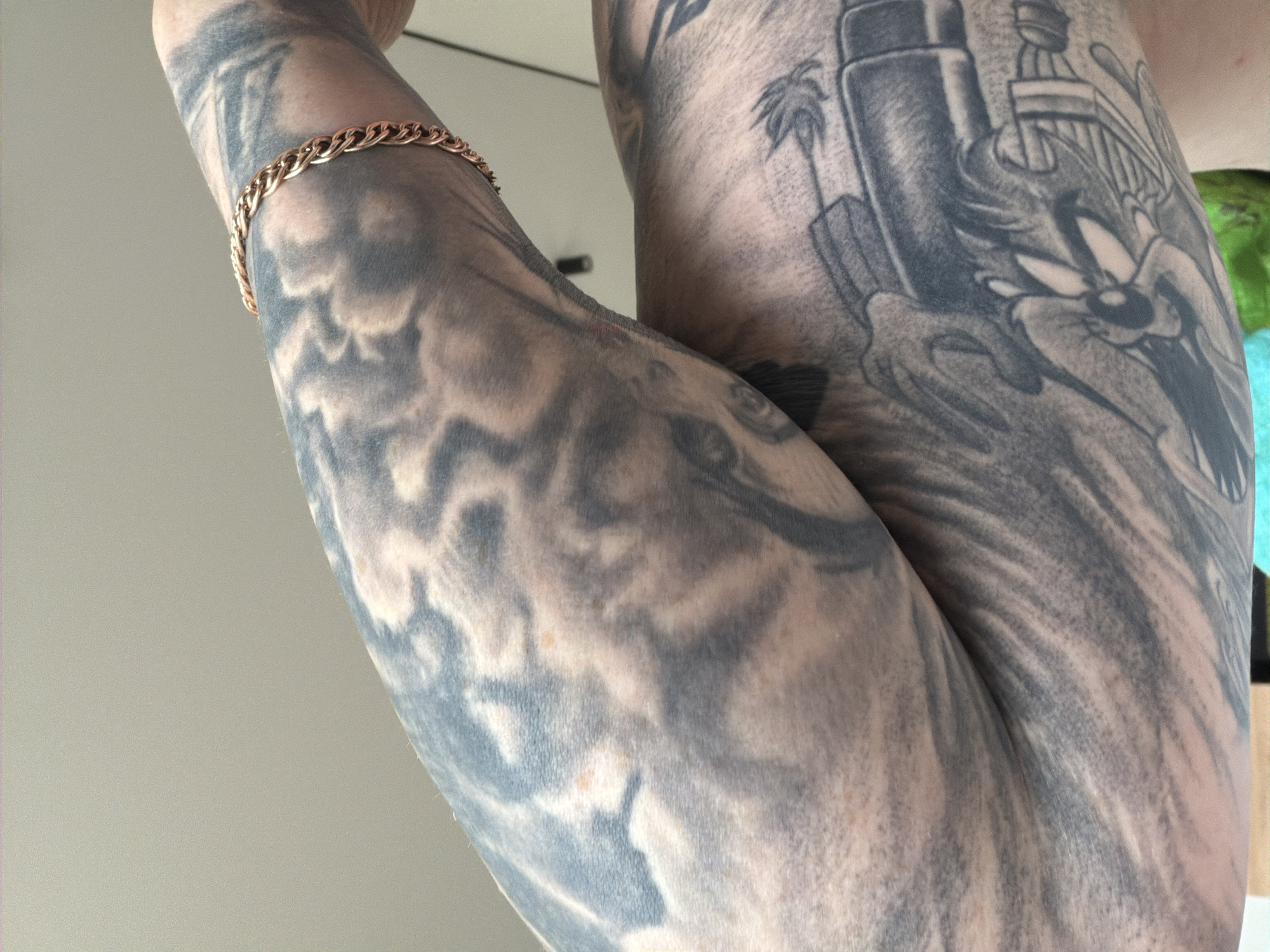 This image, captured in the city of Chaikovsky, Russia, features two human arms, heavily tattooed in monochromatic grey and black ink, positioned against an indistinct light grey background. The arm extending from the upper left displays a faded tattoo with a cloud-like or abstract pattern, showing areas of lighter skin and several small, irregular reddish-brown spots. A gold-toned, braided chain bracelet encircles this arm. The arm situated below and to the right reveals denser tattooing, including partial representations of a cartoon character resembling Wile E. Coyote, a mechanical or weapon-like design, and a palm tree illustration. Both arms exhibit visible skin texture and hair follicles.