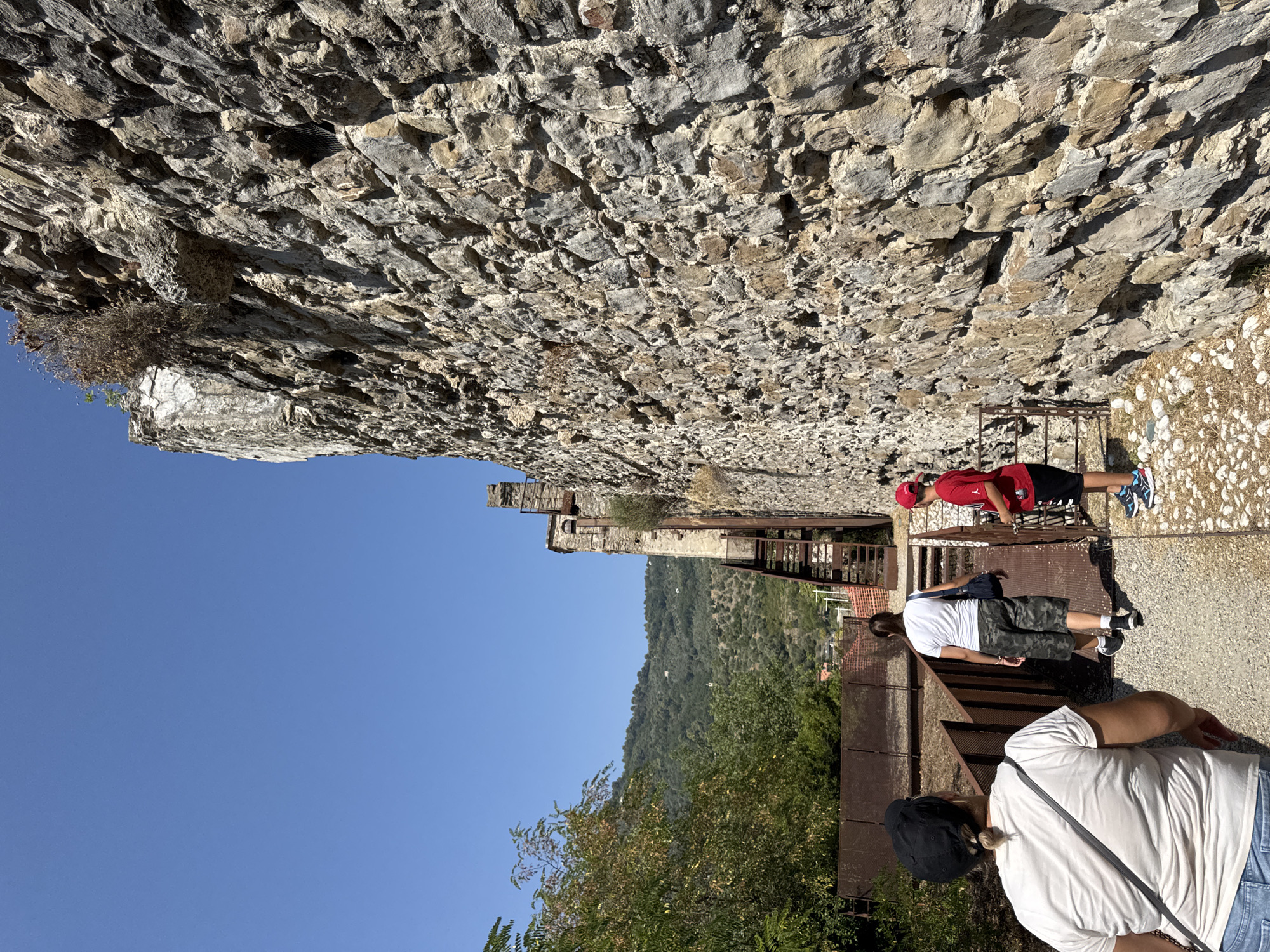 In Dolceacqua, Italy, three people are walking on a gravel path alongside a rough stone wall. The person in the foreground, viewed from behind, wears a white t-shirt and a black baseball cap. Ahead of them, a person in a white t-shirt and camouflage shorts walks away, carrying a dark blue bag. To the right, a person in a red t-shirt and black shorts, wearing a red baseball cap, stands beside a metal gate. Behind the people, a rusty metal structure with stairs ascends alongside the stone wall, and further back, a green hillside with scattered buildings and trees is visible under a clear blue sky.