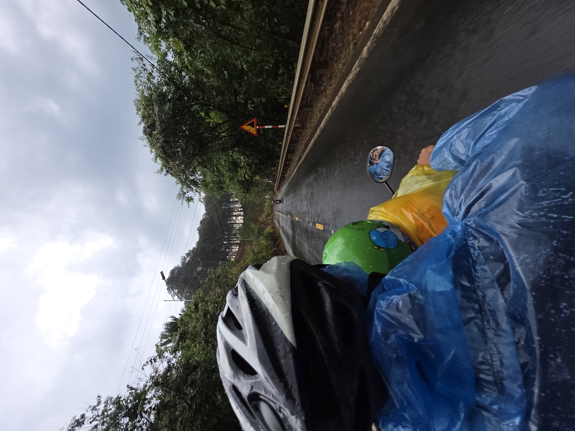 A first-person view from a vehicle, likely a motorcycle or bicycle, traveling on a wet road in Thôn 5, Vietnam. The scene is overcast, and rain is evident on the road surface. The foreground features parts of the vehicle, including a black and white helmet and yellow and blue rain gear covering items in the back. A side mirror shows a reflection of the rider. The road has a yellow centerline and a guardrail on the right. A triangular road sign is visible along the road, with trees lining both sides. The road appears to lead uphill into a forested area with glimpses of structures further in the distance.