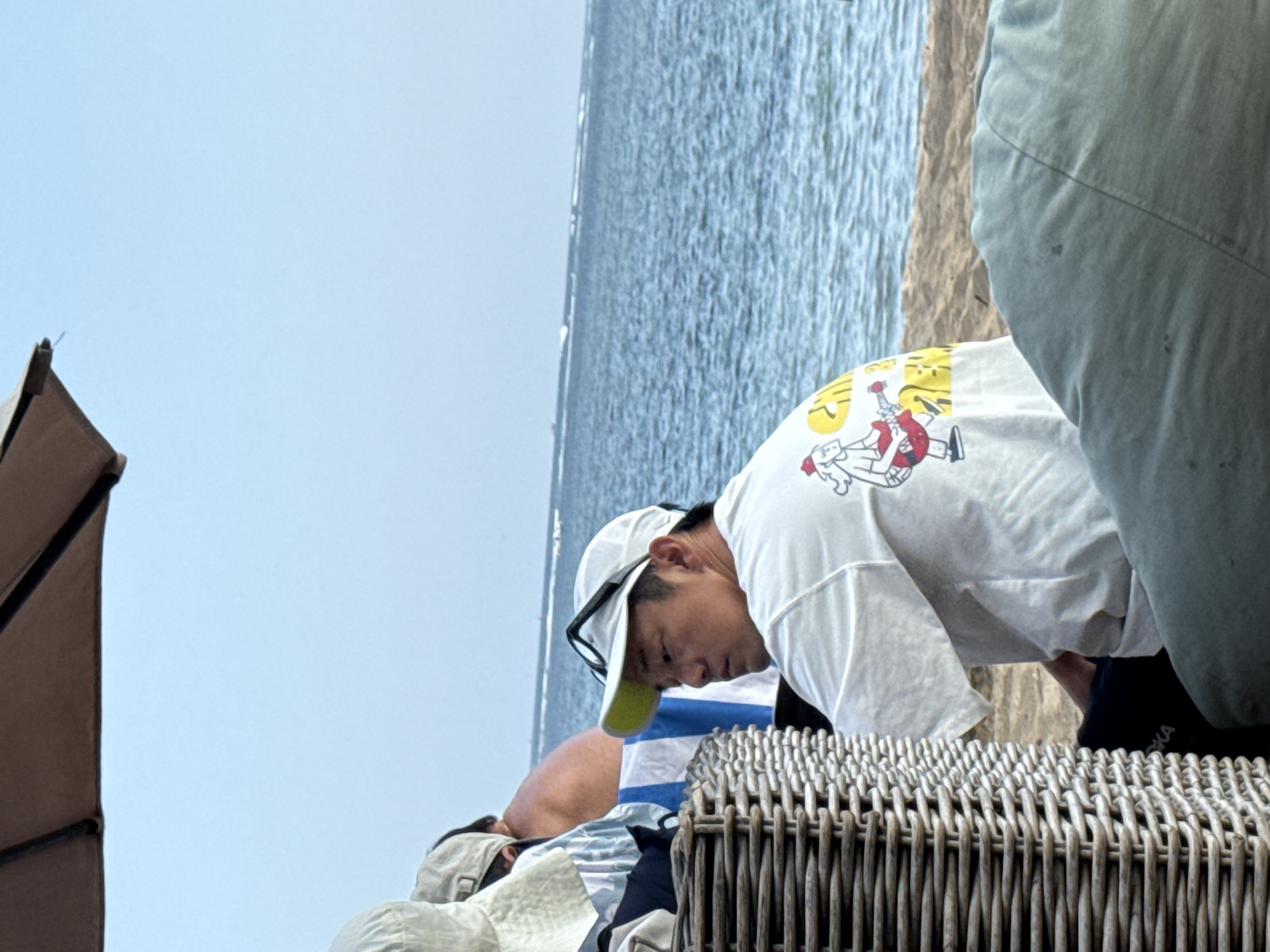 A man wearing a white t-shirt with a graphic of a cartoon character playing a guitar and a white baseball cap with a yellow brim is sitting in a wicker chair on a beach in Denpasar, Indonesia. He is looking to his right. Behind him, another person is partially visible, wearing a light-colored hat and a striped shirt. The background consists of the ocean and a clear blue sky. There is a beach umbrella visible in the upper left corner of the image.
