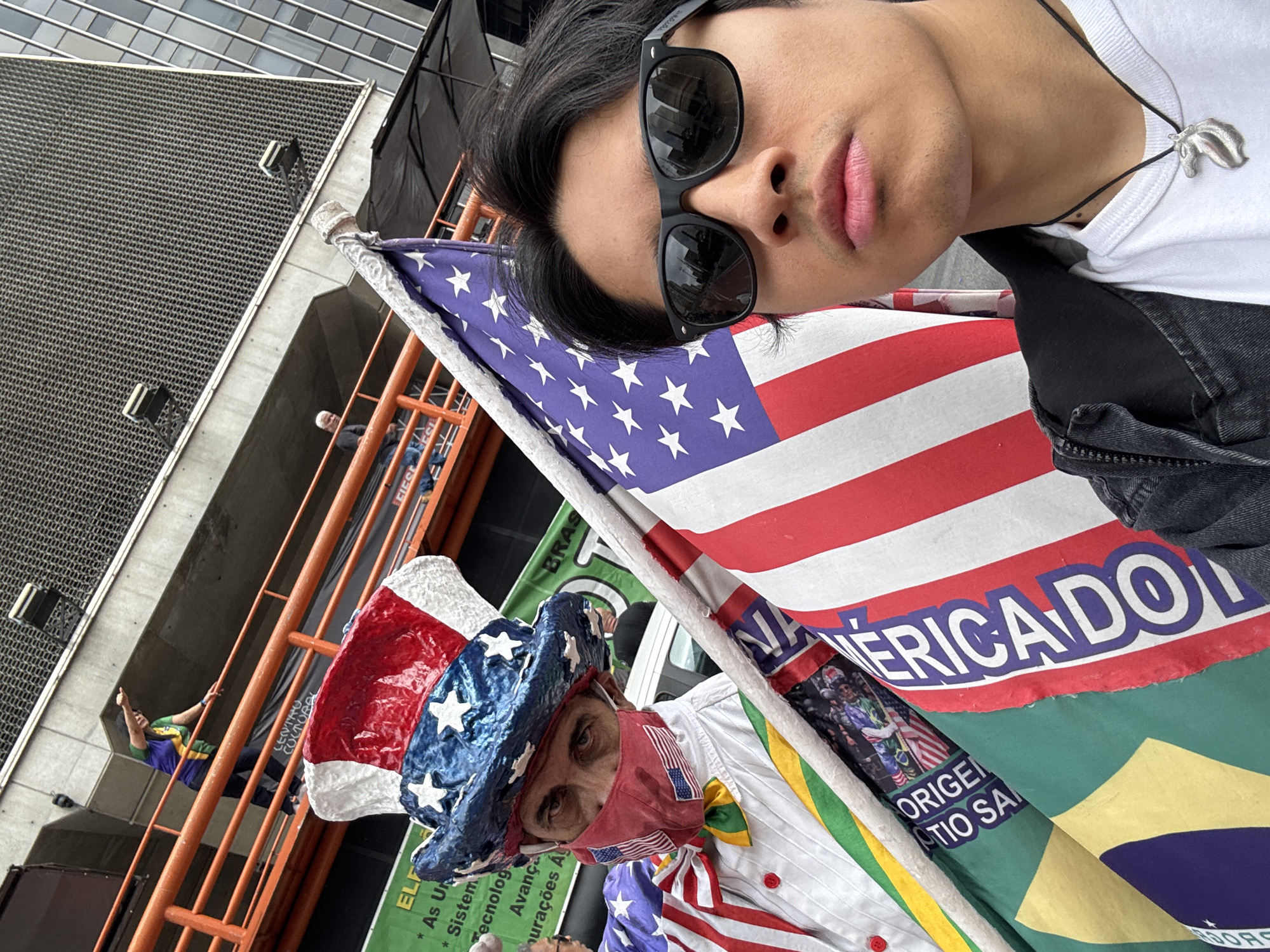 The image captures a selfie taken by a young man at an outdoor public gathering in São Paulo, Brazil. The main subject, wearing sunglasses, a white t-shirt, and a black jacket, looks directly at the camera. A silver pendant, possibly a dolphin or whale tail, hangs from his neck.
Below and to the right, another man is prominently featured, dressed in highly patriotic attire: an Uncle Sam-style hat adorned with stars and stripes, a red face mask patterned with small American flags, and a white shirt with a yellow and green bowtie. He has a serious expression.
A large American flag is draped in the foreground and mid-ground. Part of a Brazilian flag, with its green, yellow, and blue colors, is also visible in the lower right. The background features an orange metal scaffolding-like structure. On this structure, another person wearing a green, yellow, and blue shirt can be seen giving a thumbs-up. Urban buildings are also visible in the upper background.
The scene takes place during the day, suggesting a public event such as a political demonstration or rally, indicated by the prominent display of both American and Brazilian flags and the patriotic clothing. Visible text on banners and flags includes "AMERICA DO", "ORIGEM", "OTIO SA", "BRAS", and "CENTRO CULTURAL", alongside fragmented words like "ELF", "Sistem", "Tecnolog", and "Avança".