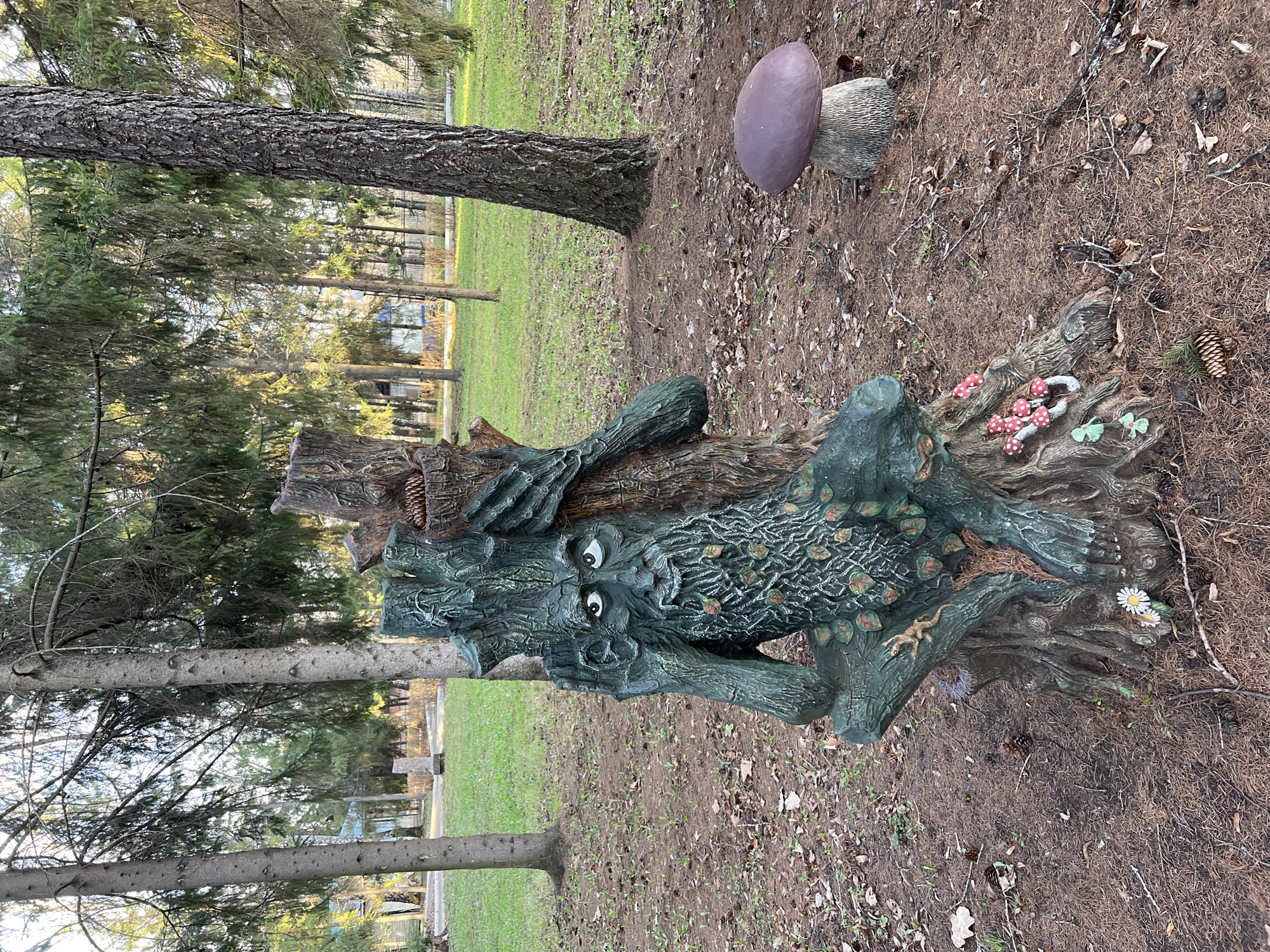 The image shows a park scene in Sanatoriya Yumatovo Imeni 15-Letiya Bassr, Russia. The focal point is a decorative tree stump sculpture with a face carved into it. The stump is painted green and features small details such as leaves and a lizard. Next to it is a mushroom statue. The ground is covered with pine needles, small twigs, and a few pine cones. Several trees surround the area, suggesting a wooded or park-like setting. In the background, a glimpse of a building or structure can be seen through the trees.