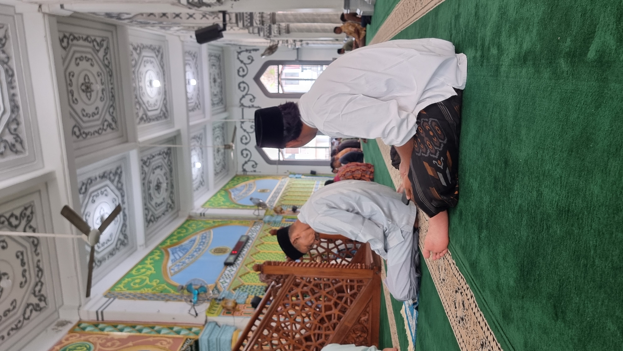 The image depicts the interior of a mosque in Keudekruenggeukueh, Indonesia, with a focus on the green carpeted prayer hall. Two men are prominently featured, both wearing traditional attire and black peci hats. The man in the foreground is seen from the rear, wearing a white long-sleeved shirt and a patterned sarong, with his legs folded. To his left, another man is in prayer, kneeling with his head bowed, wearing a light-colored robe and pants. In the background, several other individuals are also seated or in prayer, and the mosque's architecture is visible, including a wooden screen, ornate ceiling with decorative panels and a fan, and colorful wall murals. The overall atmosphere appears to be one of quiet devotion.