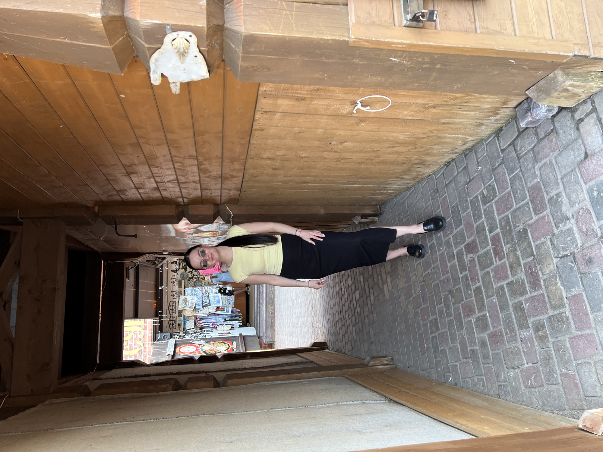 A street scene in Sviyazhsk, Russia, shows a woman wearing sunglasses, a light yellow shirt, and a black skirt, standing on a brick-paved area. Overhead, a wooden structure with carved details and a small metal owl ornament is visible. The perspective suggests she is in a narrow alley or passageway with wooden beams above. In the background, a dimly lit opening reveals a souvenir shop.