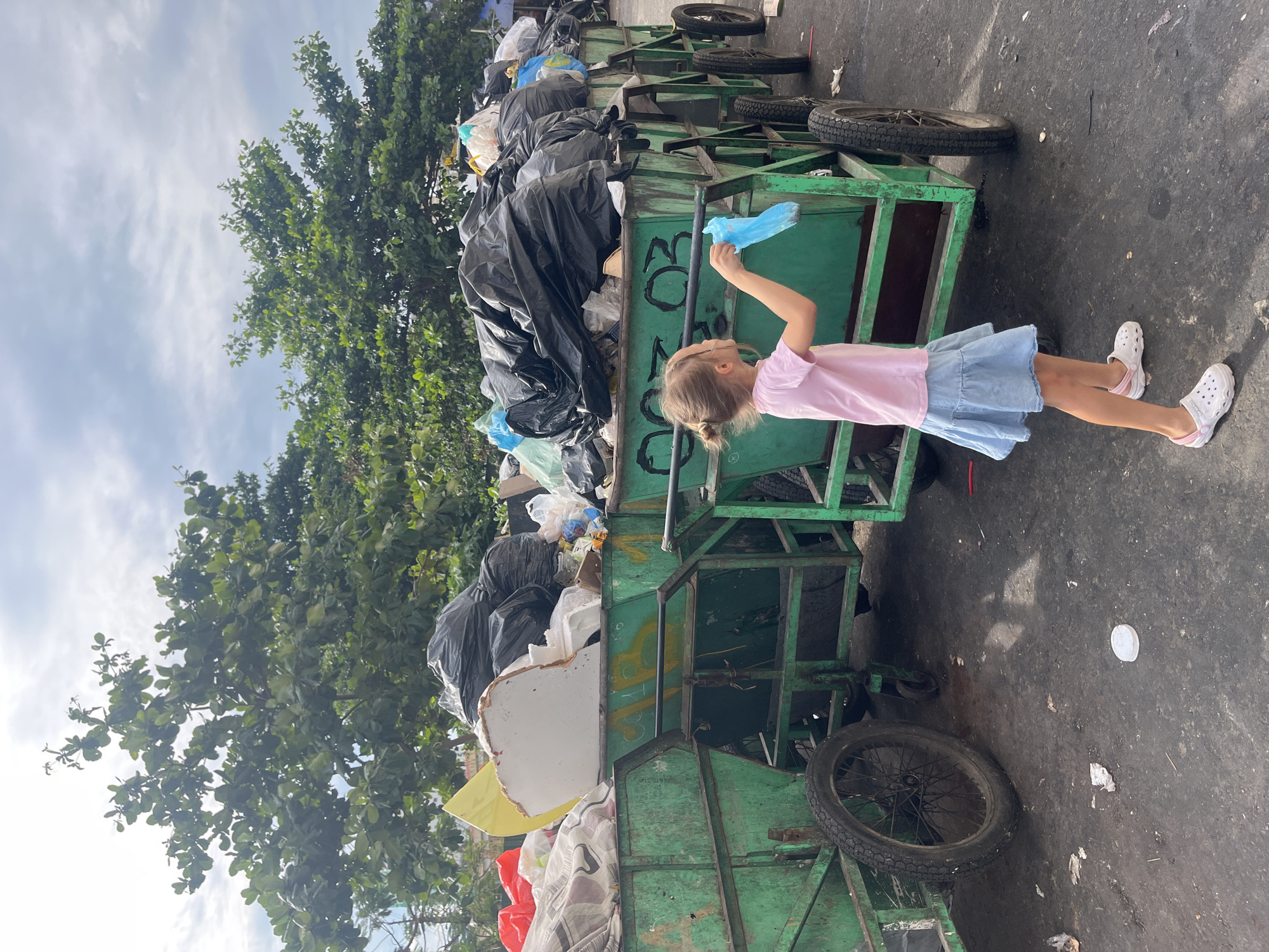 A young girl stands near several green, wheeled garbage bins filled with bags of trash. She is wearing a pink shirt, a blue denim skirt, and white Crocs-style shoes with pink details. She is holding a blue plastic bag. The bins are overflowing with black garbage bags and other types of waste. The scene is on a paved surface, possibly a street, with a large tree in the background. The sky is visible above the tree. The location is identified as Bac Nha Trang Ward, Vietnam.