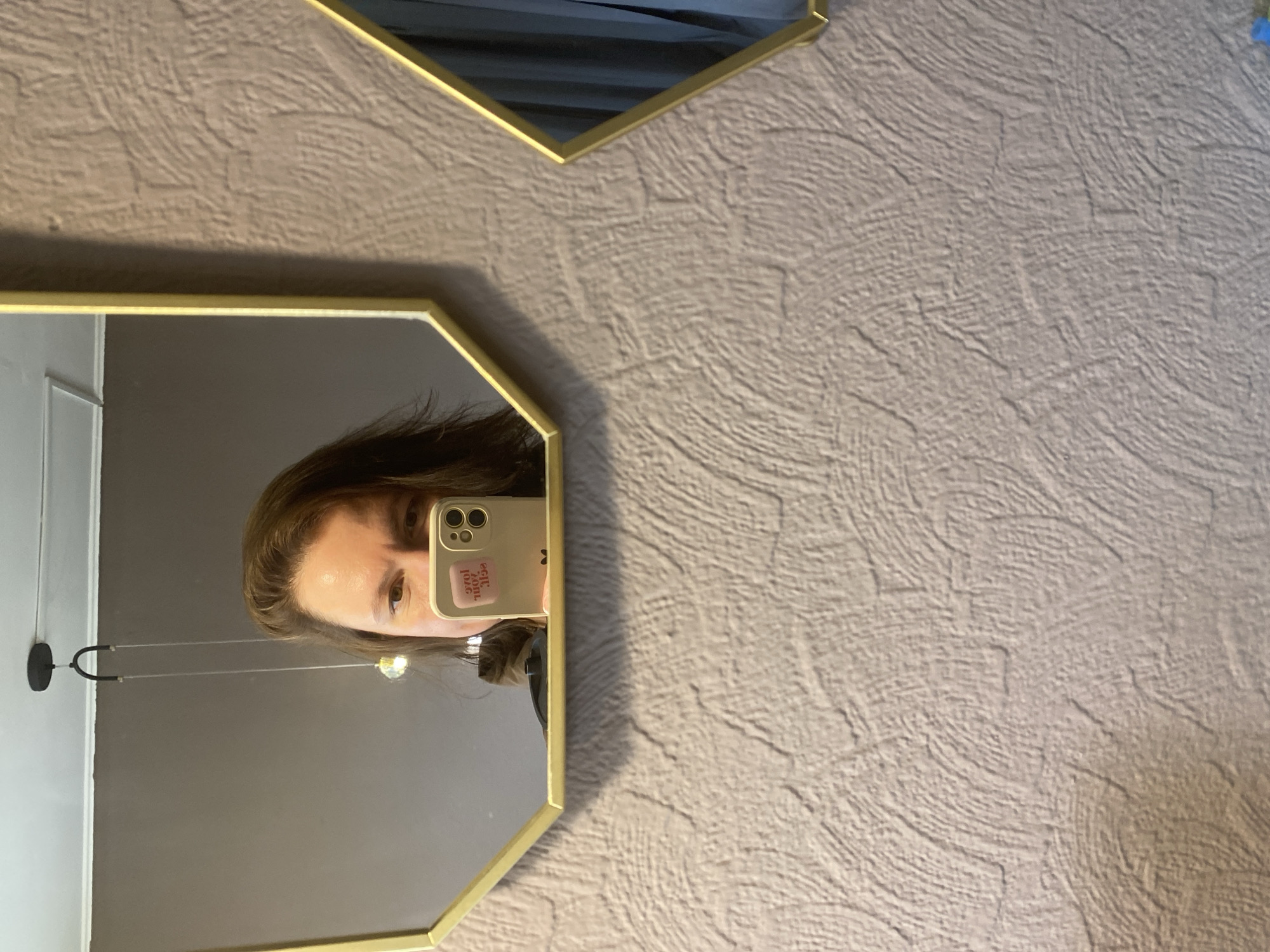 The image displays a person taking a mirror selfie in an indoor setting, likely in Chaikovsky, Russia. The central subject is the reflection of a person's upper face, including their eyes, forehead, and part of their brown hair, visible within a gold-framed hexagonal mirror. The person is holding a light-colored smartphone to their face, capturing the reflection.
On the back of the smartphone, there is a light pink sticker with visible text that appears reversed due to the mirror image. Distinct parts of the text visible include "эгл" and "200", suggesting a phrase like "2000s girl" or similar when unreversed.
The mirror also reflects a dark-colored wall, a white ceiling, and a dark, modern-looking light fixture hanging from the ceiling. A second, partially visible gold-framed hexagonal mirror is situated above the main one, reflecting a dark, indistinct area. The wall upon which the mirrors are mounted is covered with textured, light beige or off-white wallpaper featuring a subtle, wavy pattern. The overall impression is an interior space with contemporary decor. The lighting suggests daytime or a well-lit indoor environment. No specific emotions are discernible from the person's neutral expression.