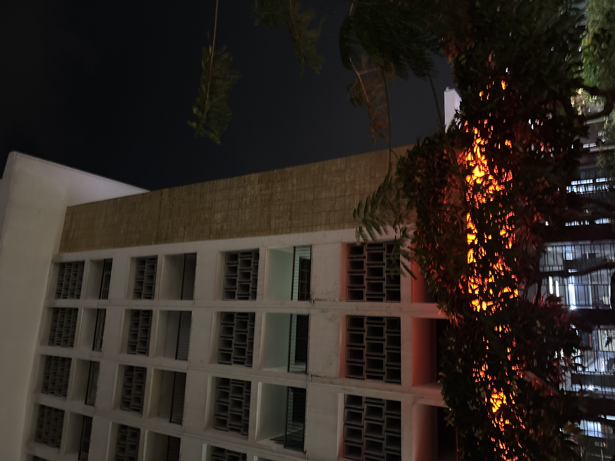 An exterior, low-angle night shot captures a stark, Brutalist-style building with a light-colored facade featuring rows of recessed windows and balconies. A vertical, textured concrete pillar extends upwards along the right side of the building. In the foreground, lush green trees are illuminated from below by a vibrant orange-red light, creating a fiery effect on their leaves. The dark night sky is visible behind the trees and building. The building is identified as the Central Bank of Indonesia (Bank Indonesia) in West Jakarta, Indonesia. No people are visible in the image.