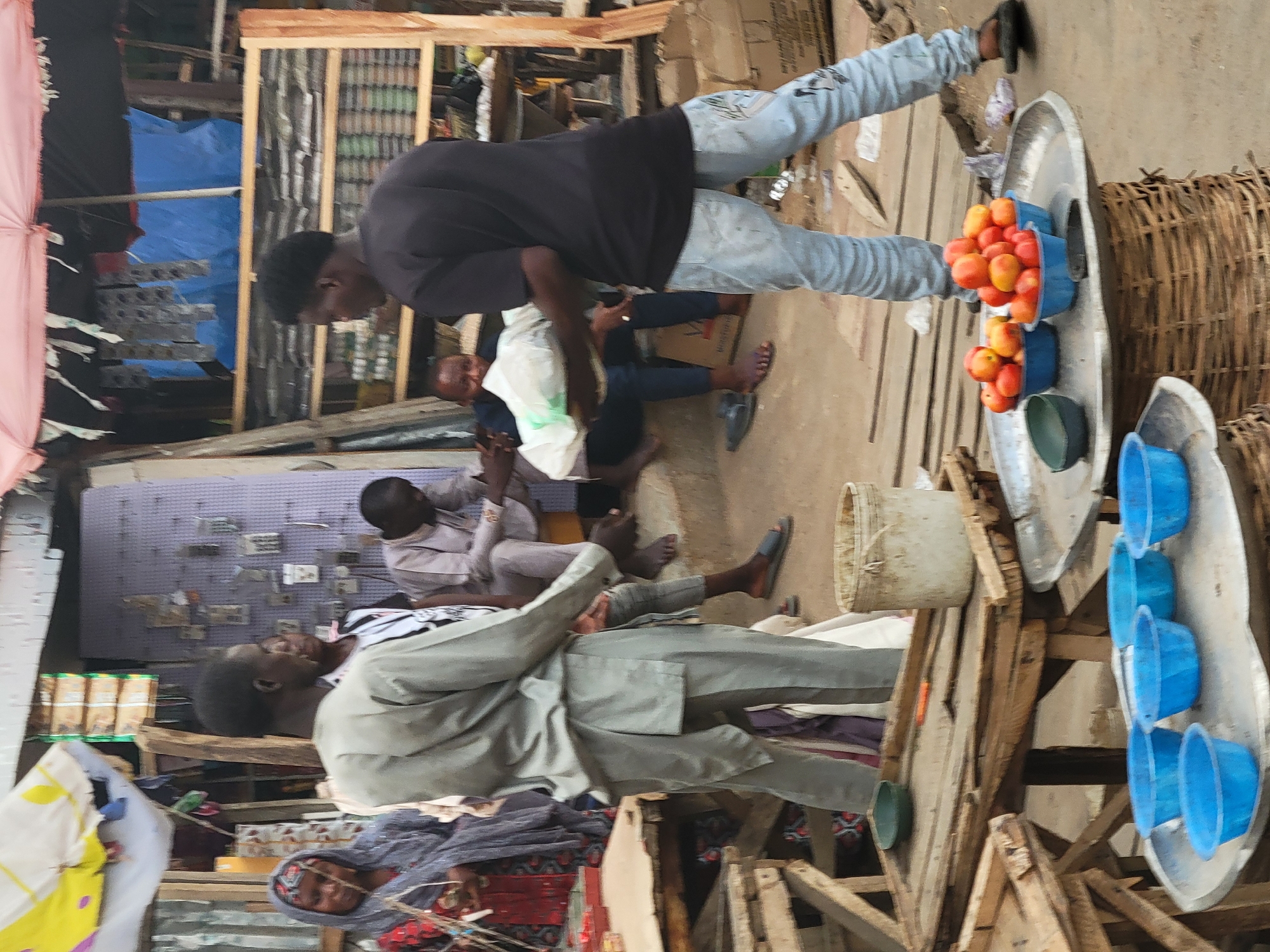 The image depicts a bustling scene in Abuja, Nigeria. Several people are visible in a market environment, with a man in a grey traditional outfit seated and conversing with another man. Further in the background, other individuals are seated, possibly engaging in conversation or observing the surroundings. To the right, a vendor is attending to a display of goods, including what appear to be apples in blue containers on a large silver tray. A variety of containers, potentially for selling goods, are seen adjacent to the vendor. The environment appears to be crowded with market stalls and activity.
