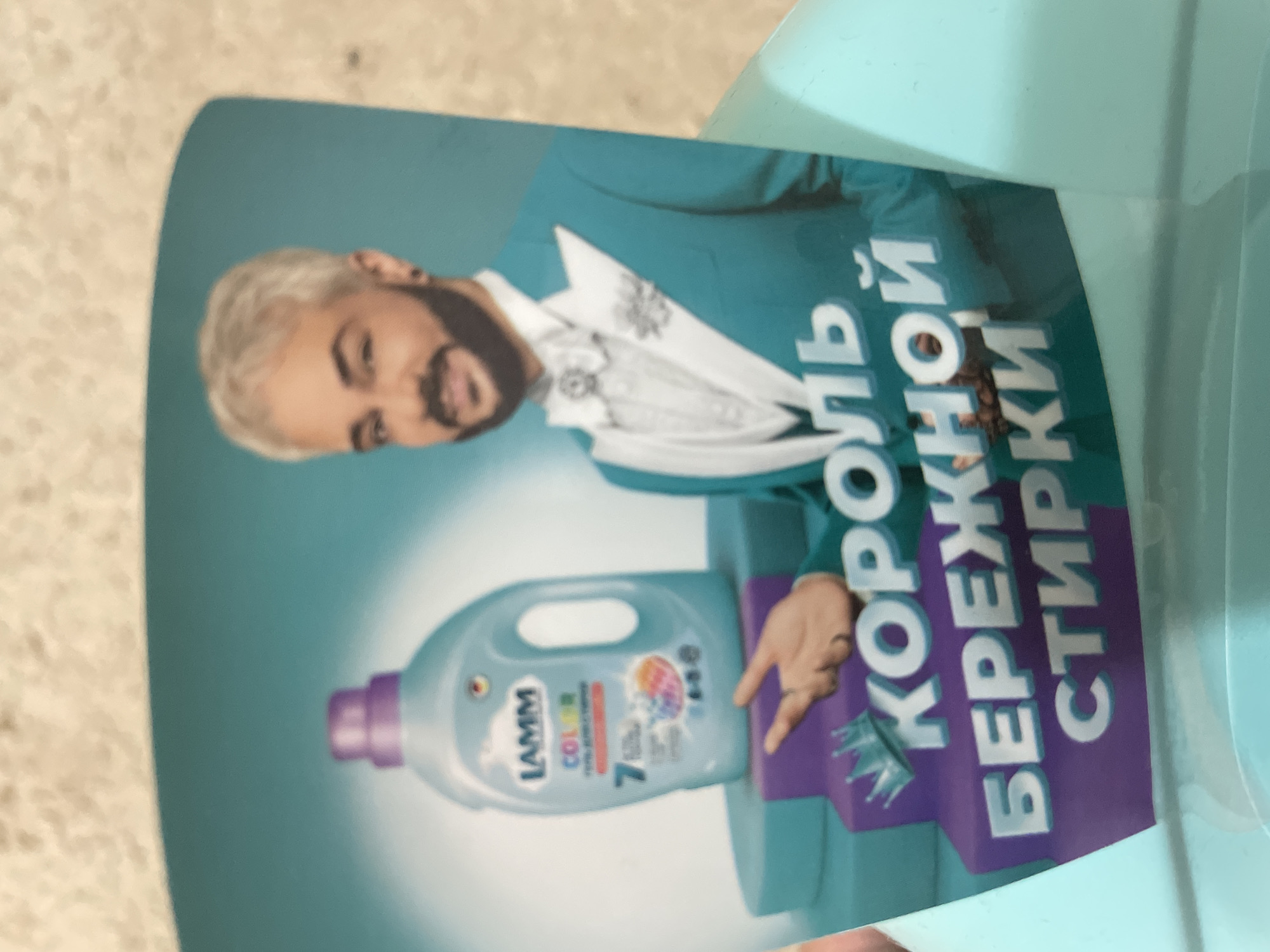 The image is a close-up photograph of a printed advertisement for LAMM COLOR laundry detergent, observed in the city of Votkinsk, Russia. The specific geographical coordinates or context within Votkinsk are not discernible from the visual content of the image itself.
The advertisement features a male figure standing to the right of a bottle of detergent. The man has light-colored hair, a neatly trimmed dark beard and mustache, and dark eyebrows. He is smiling with his mouth closed. He wears a light teal suit jacket with white lapels, a white waistcoat, a white collared shirt, and a white, ornate bow tie. A decorative brooch is pinned to his left lapel. A dark earring is visible in his left ear. His right hand is extended, palm slightly up, gesturing towards the detergent bottle. A ring is visible on his right ring finger.
To the left of the man, a light blue LAMM COLOR laundry detergent bottle with a purple cap and handle rests on a three-tiered platform. The top tier is teal, and the bottom two tiers are purple. The bottle's label clearly shows "LAMM" and "COLOR" text, along with a small German flag icon, the number "7", and an indistinct colorful graphic.
Large, white Cyrillic text is displayed on the purple tiers of the platform. On the upper purple tier, a silver crown icon precedes the word "КОРОЛЬ" (King). Below this, on the next tier, is "БЕРЕЖНОЙ" (Gentle/Careful). The lowest visible text on the bottom tier is "СТИРКИ" (Washing/Laundry). The full phrase translates to "KING OF GENTLE WASHING".
The advertisement's background is a plain, smooth teal color. The photograph itself shows the printed advertisement resting against a textured, light-colored surface at the top, possibly a wall, and positioned within a light blue, smooth, curved object visible at the bottom and right edges of the frame.