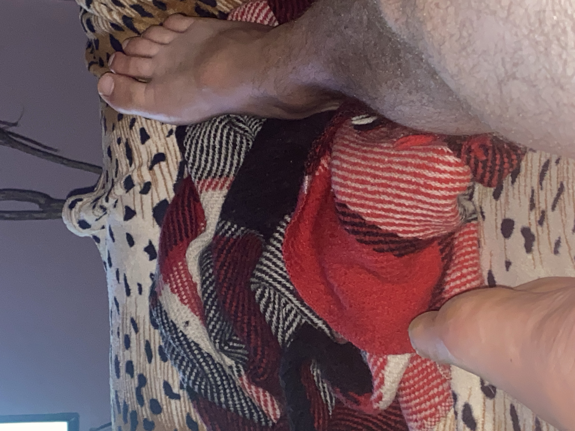 The media file shows a close-up of a person's feet resting on a patterned blanket. The blanket has a leopard print and a red, black, and white plaid design. The person's legs are partially visible, with one leg covered in hair. The scene appears to be indoors, with a dim purple background suggesting it is nighttime or the room is dimly lit. There are no visible text elements in the image. The focus is on the textures of the blanket and the skin of the feet, creating an intimate and relaxed atmosphere. The overall impression is one of comfort and repose.