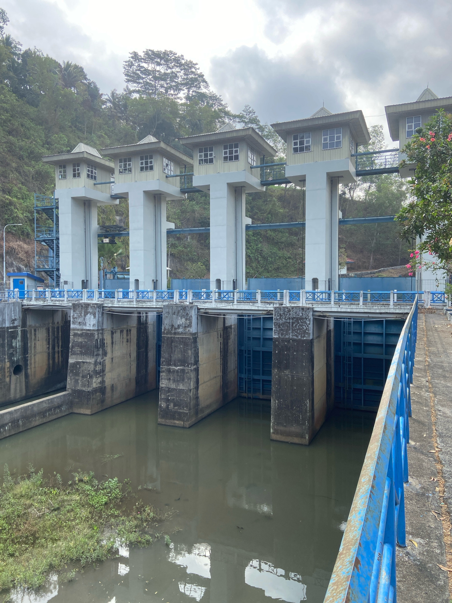 The image shows a water dam with multiple gates and control towers. The dam structure is made of concrete and steel, with a blue railing along the walkway. The water in front of the dam is murky and has some vegetation growing in it. The background shows a lush green hillside covered with trees. The sky is overcast with clouds. The dam is likely located in Segawe, Indonesia, given the context. There are no people visible in the image.