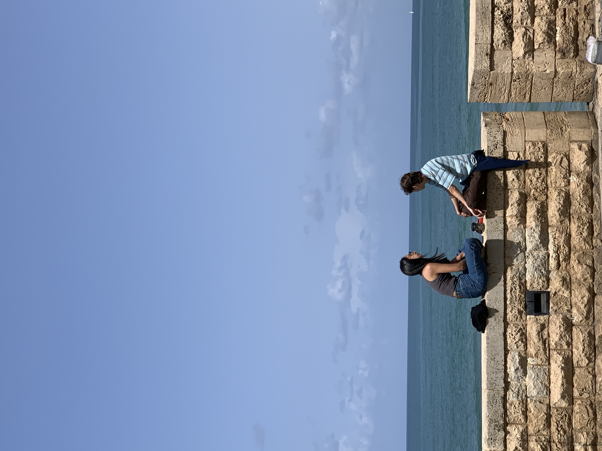 Two individuals are positioned on a stone wall overlooking the ocean. A male, wearing a striped shirt and dark pants, is leaning towards the other. A female, wearing a tank top and jeans, is seated. The backdrop includes the ocean and a partly cloudy sky. The setting is likely a part of the historic city of Jaffa, now incorporated into Tel Aviv, Israel, given the architectural style of the stone wall.
