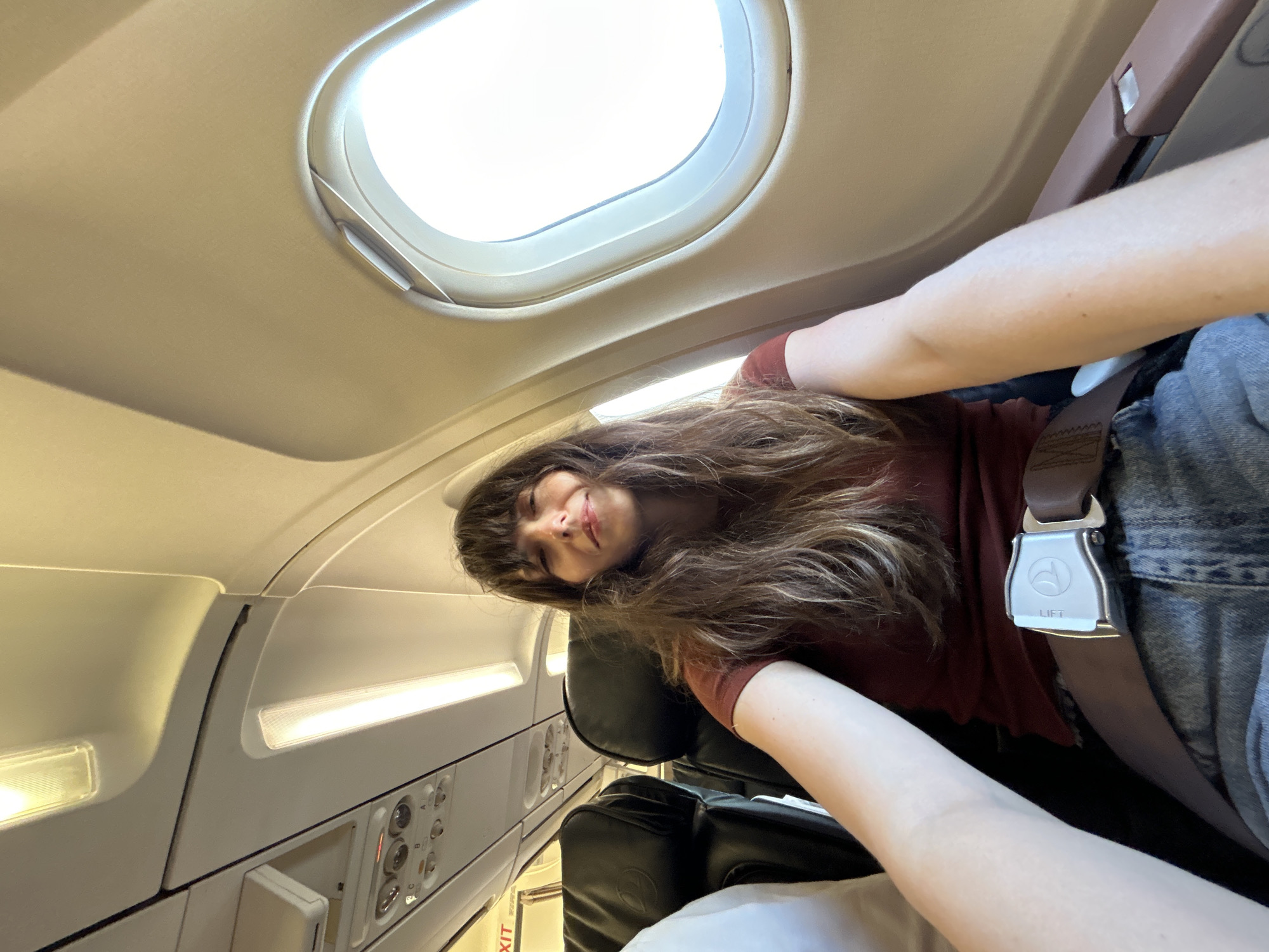A woman with long, dark, wavy hair and bangs is seated in a plane. She is wearing a maroon shirt and denim jeans, with her seatbelt fastened. The seatbelt buckle is silver with a "THY" logo. To her left are two black airplane seats, one occupied by an unseen person, and to her right is an airplane window. The interior of the plane is visible, including overhead lights and an "EXIT" sign. The scene takes place inside an airplane, likely at Dalaman Airport in Turkey, based on the "THY" logo which is the abbreviation for Turkish Airlines.
