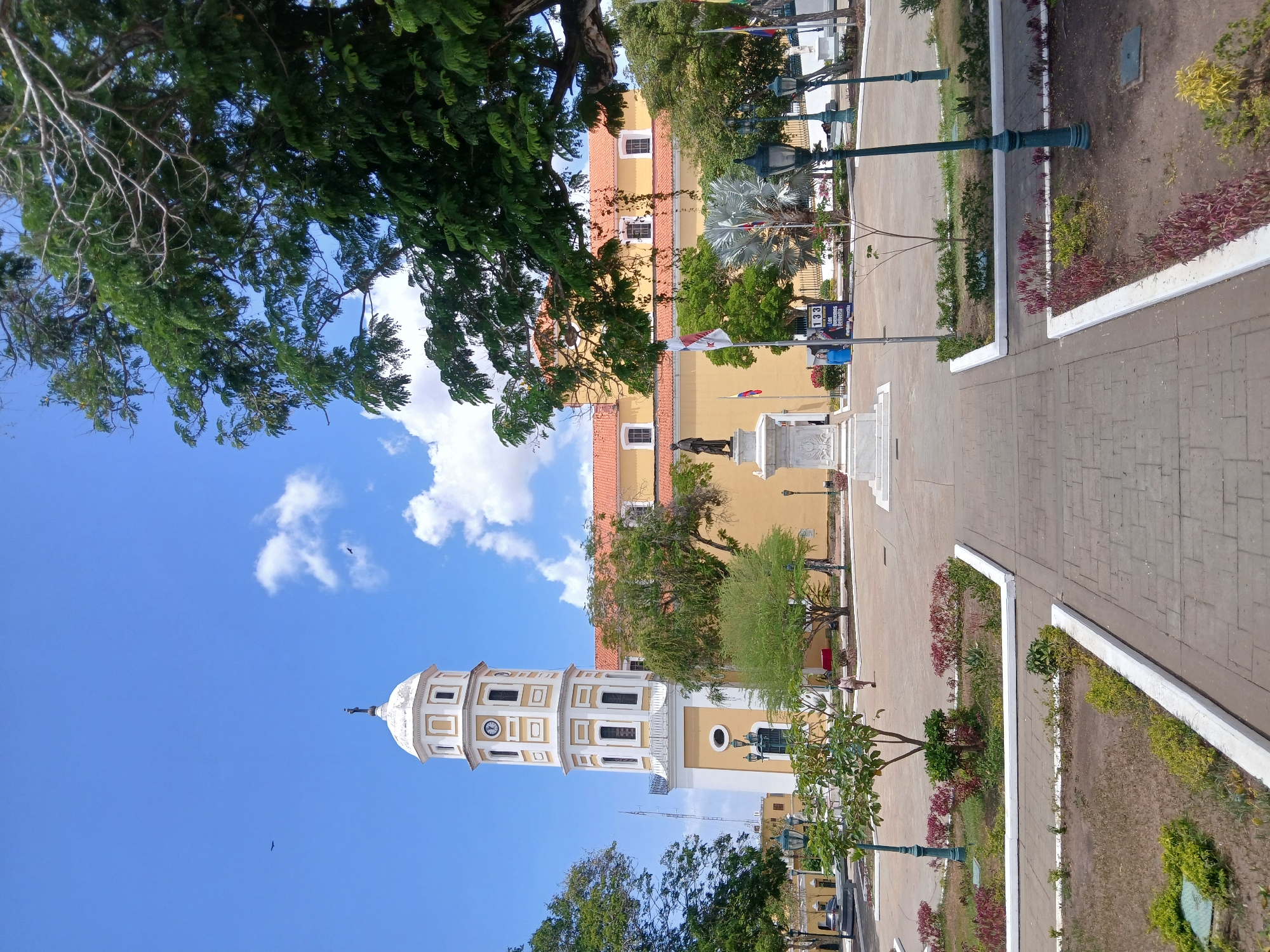 The image captures a scene in a public square in an unknown city. A white and yellow bell tower with a clock stands prominently, adjacent to a larger yellow building, likely a church. A statue on a pedestal is centered within the square. People are seen walking around the square. The square is paved with stone, and there are landscaped areas with flowers and greenery, bordered by white painted edges. Green street lamps are interspersed throughout the square. The sky is blue with scattered white clouds. Trees are visible around the perimeter of the square. Flags are visible in the background.