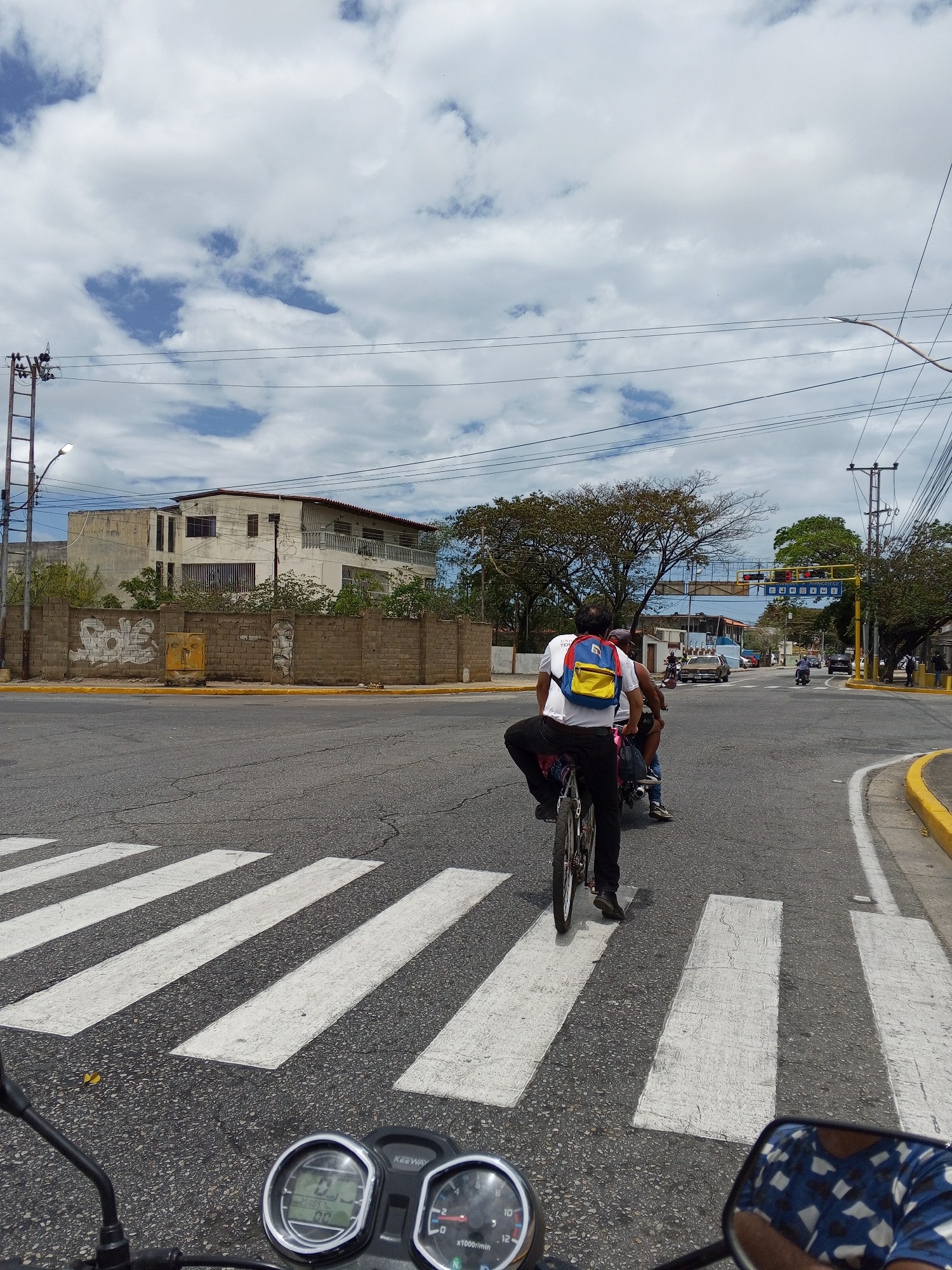 The image depicts a street scene in Juan Griego, Venezuela. The view is from the perspective of a motorcycle rider stopped at a crosswalk. A man on a bicycle is crossing the street in front of the motorcycle, carrying a passenger on the back. The cyclist is wearing a white shirt, black pants, and a backpack with red, blue, and yellow sections. Other vehicles, including motorcycles and cars, are visible in the background. Buildings and trees line the street. The sky is partly cloudy. Street signs are visible, and power lines run overhead.