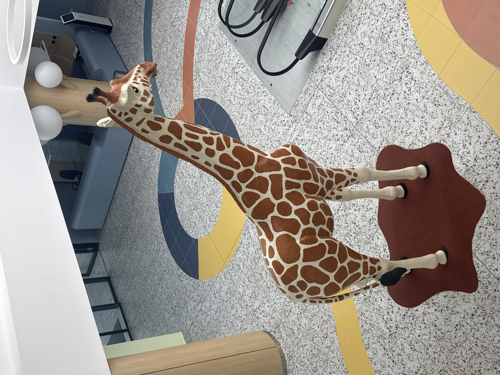 The image shows a large, life-sized giraffe statue positioned in the center of a modern, open-plan interior space. The floor is a speckled terrazzo with colorful, curved geometric patterns in yellow, blue, and coral. To the right of the giraffe, an escalator is visible, with its metal treads and handrails. In the background, there is a reception desk with blue seating, office chairs, and a digital kiosk. The overall environment appears to be a public building, possibly a mall, office, or educational institution. The giraffe statue is the primary subject, its brown and white pattern clearly defined. The lighting suggests an indoor, well-lit space. The scene is static, with no visible people or activity. The image does not provide any specific information about the city of Burgas, Bulgaria.