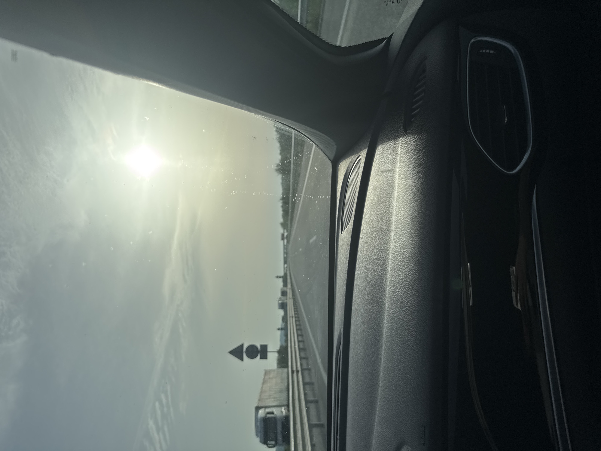 The image shows a view from inside a car on a highway. The sun is visible through the windshield, causing glare. There is a truck and a direction sign on the side of the road. The interior of the car is visible, including part of the dashboard and air vent. The location is unknown, but it has nothing to do with Arnautito, Bulgaria.