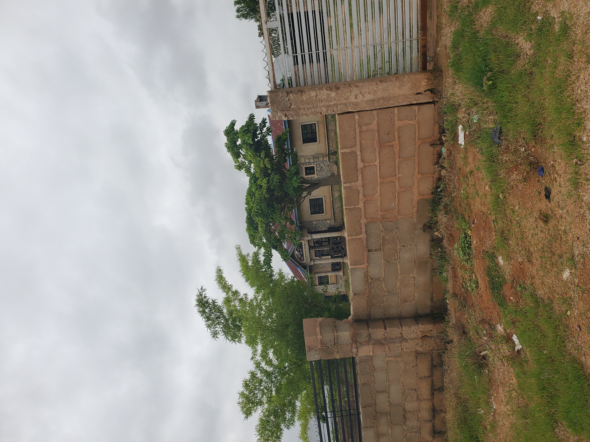 A low-angle outdoor medium shot depicts a house and its surrounding fence in Du, Nigeria, under an overcast sky. The house, a single-story structure with a red corrugated metal roof, is partially visible behind a substantial fence. The fence is constructed from two types of materials: a lower section made of light brown bricks, approximately waist-high, topped with a dark metal railing, and a taller, light gray metal gate on the right side of the frame. The house itself features light-colored walls with stone accents around the windows and entrance. A portion of the entrance is visible, showing classical-style columns supporting a pediment. Two trees are present: a large bamboo-like plant with light green foliage to the left of the house, and a leafy green tree with a rounded canopy in front of the house. The ground in the foreground is covered with dry grass and dirt, with some scattered debris, including plastic bags. The overall impression is of a residential property under construction or a recently completed home in a somewhat undeveloped area.