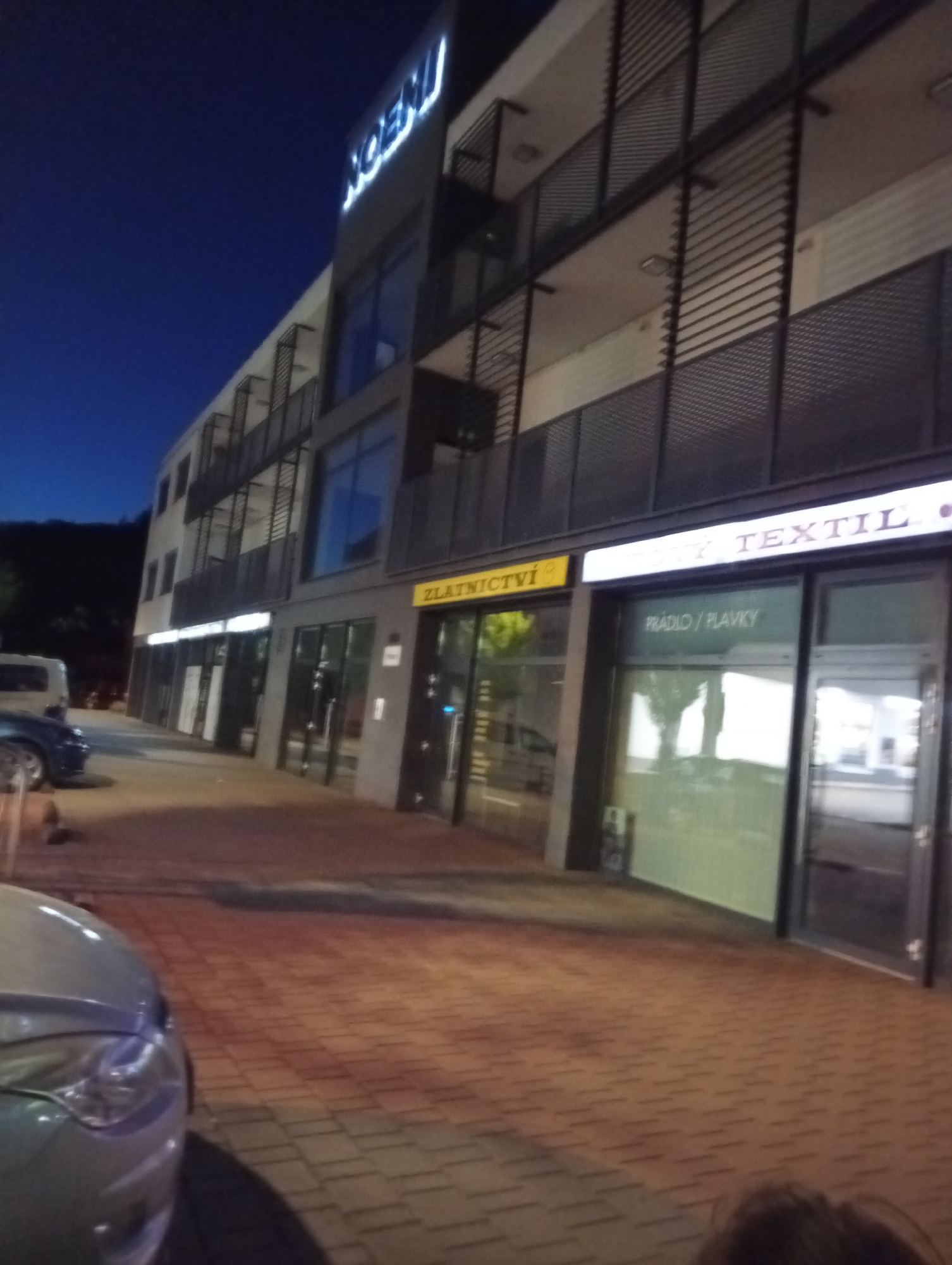 This is a nighttime outdoor image of a modern building with businesses on the ground floor. The building appears to be a multi-story structure with balconies and contemporary architectural elements. A prominent sign with blue neon letters that reads "NOEMI" is visible at the top of the building. On the ground floor, there are storefronts with illuminated signs. One sign is yellow and reads "ZLATNICTVI", likely indicating a jewelry store. Another sign above a storefront reads "TEXTIL". Below the "TEXTIL" sign, a smaller sign within the window displays "RÁDIO / PLAVKY". The street in front of the building is paved with brick-like stones. A silver car is partially visible in the foreground on the left, and a white van is parked further down the street on the left. The sky is dark blue, suggesting it is evening or night. The overall environment appears to be a commercial area. The building and businesses are located in Tišnov, Czechia.