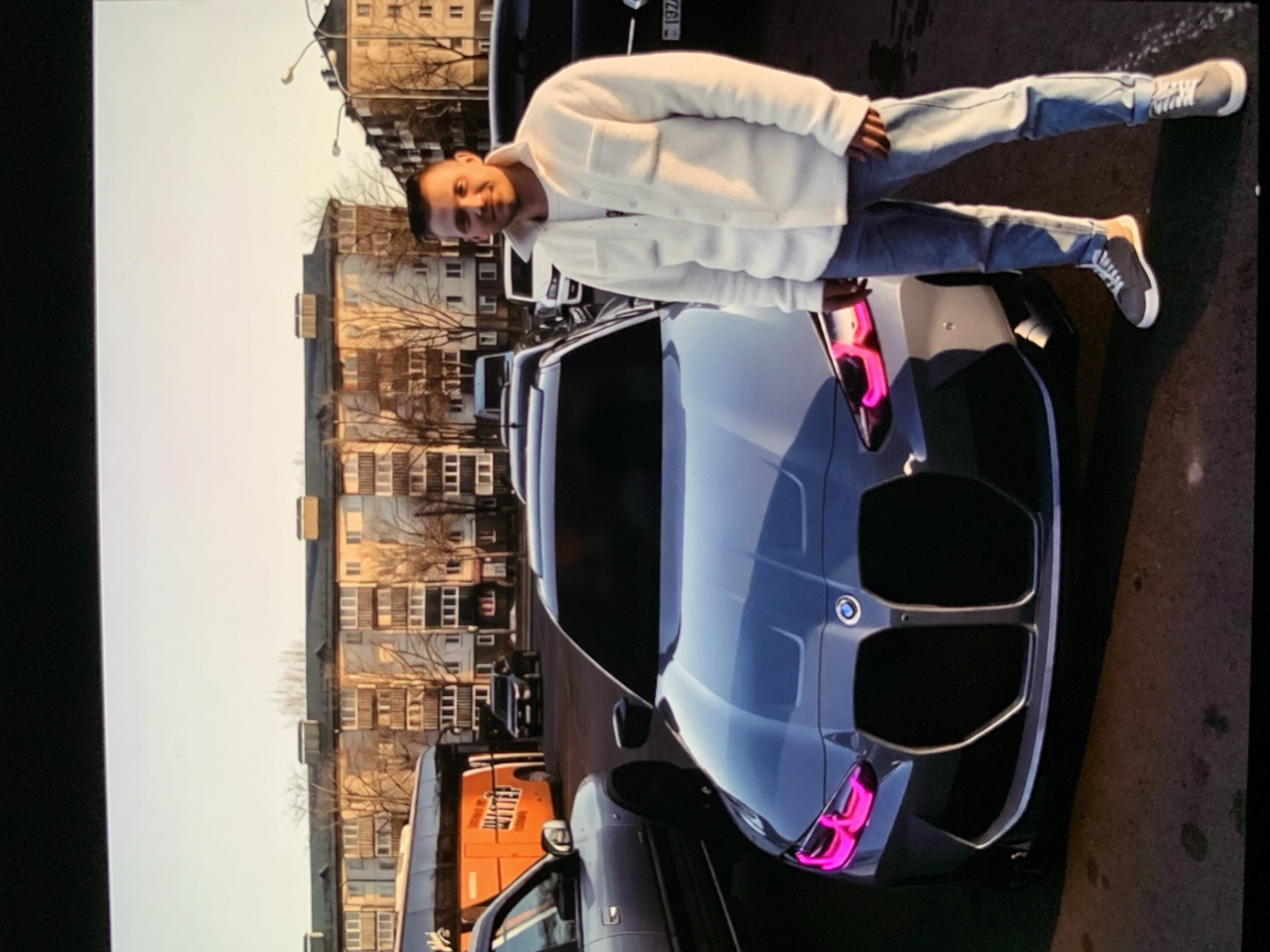 A man stands in front of a modern gray BMW with pink accents on the headlights, in what appears to be a parking lot in front of apartment buildings. The man is wearing a white fleece shirt over a white t-shirt, and blue jeans. He is looking at the camera and smiling. In the background, there are several other vehicles, including a bus and a white van, and multi-story apartment buildings. The sun appears to be low in the sky, casting long shadows. The overall scene is urban and casual.
