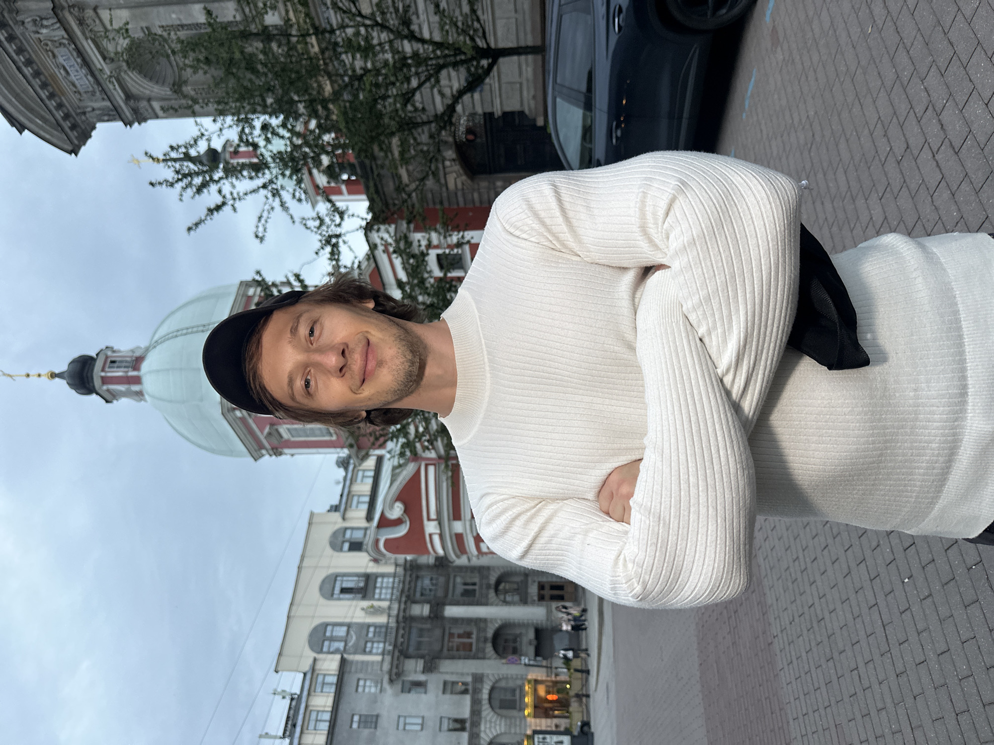 A medium shot captures a young adult male with light brown hair and a beard standing with his arms crossed in front of him. He is wearing a white, ribbed, long-sleeved sweater and a black baseball cap. He smiles slightly at the camera.
The man is positioned outdoors in a city square with a cobblestone pavement. In the background, several buildings are visible. To the left, a multi-story building with an arched entrance and numerous windows is seen. To the right, a domed building with a red and white facade, topped with a cross, stands prominently. Adjacent to this, another building with a grey, ornate facade and an arched entrance is visible, with trees partially obscuring it. A dark-colored SUV is parked to the right of the frame. The sky is overcast, suggesting it is either early morning or late evening.
The overall scene depicts an urban setting, likely in Saint Petersburg, Russia, given the architectural style of the buildings, particularly the domed structure which resembles religious architecture common in the city. The man appears to be a tourist or resident enjoying a moment in the city.