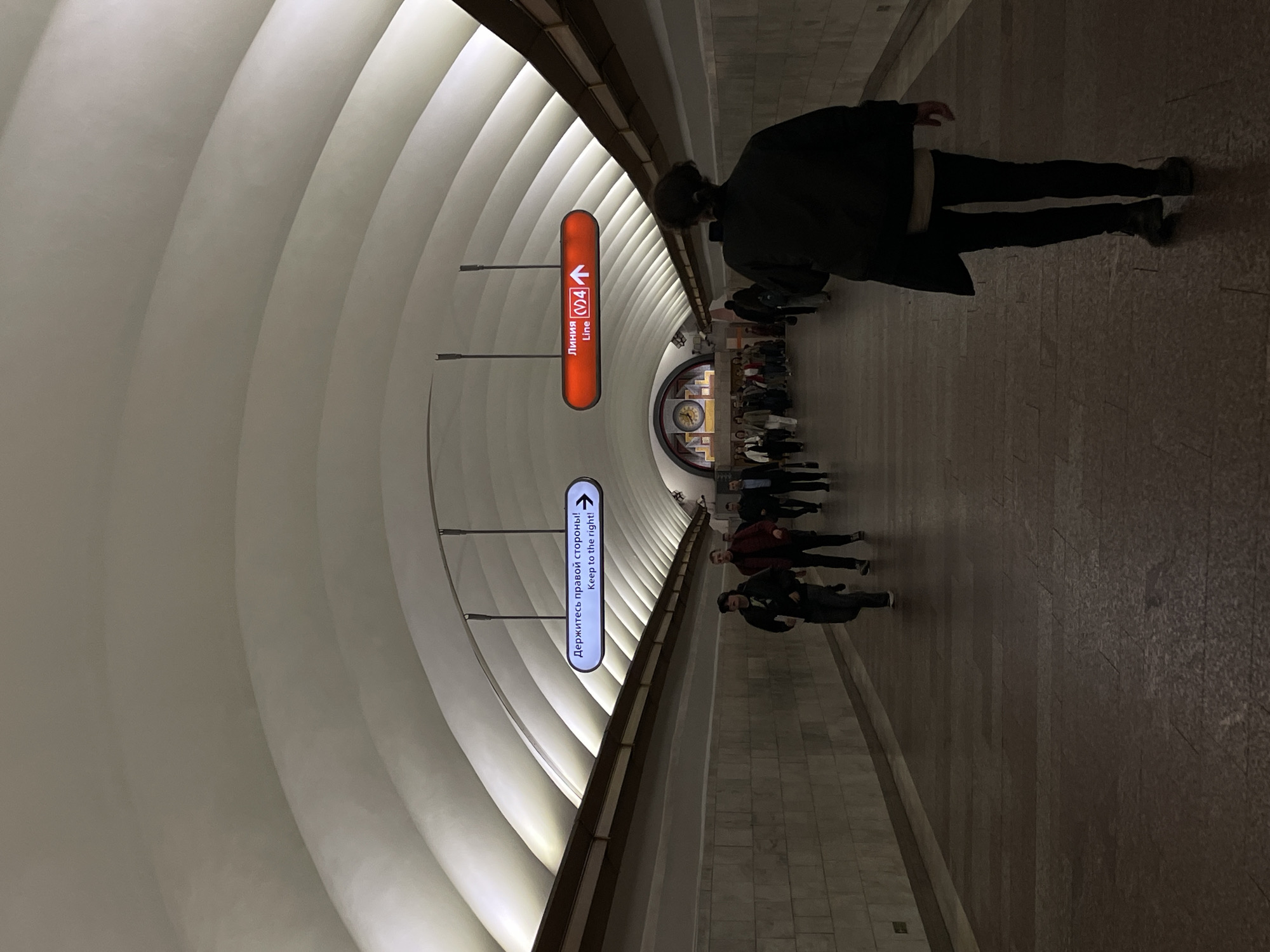 Here is a detailed description of the image:
The image captures the interior of a subway station, characterized by a long, arched ceiling with repetitive ribbed patterns. Numerous people walk along the walkway, suggesting transit within the station. Two signs, one red and one blue, are suspended from the ceiling, featuring directional arrows and text in both Russian and English: the red sign is for the "Line 4" and the blue one says "Keep to the right!".
The floor and walls are finished with a textured material, possibly stone or tile. Lighting appears diffused, illuminating the station while maintaining a relatively subdued tone. The foreground features a person walking, obscuring some of the background detail, but still, people are visible in the distance, congregating near what appears to be a clock or decorative element at the end of the walkway.
Based on the presence of Russian text alongside English translations, the station is likely located in a city with a significant Russian-speaking population, although the exact city is unknown.