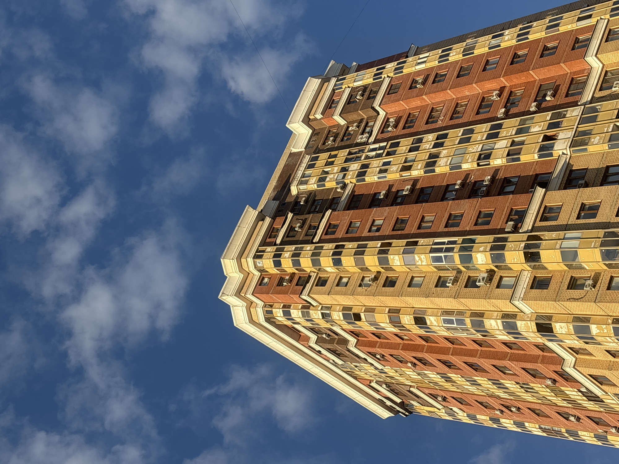 A tall, modern apartment building with a multi-colored facade of red and yellow brick and numerous windows stands against a bright blue sky with scattered white clouds. The building's top is adorned with ornate white trim. Several air conditioning units are visible mounted on the exterior walls. The perspective is looking up at the building from a low angle. The image does not contain any people, actions, or interactions. The location is Moskovskiy, Russia.