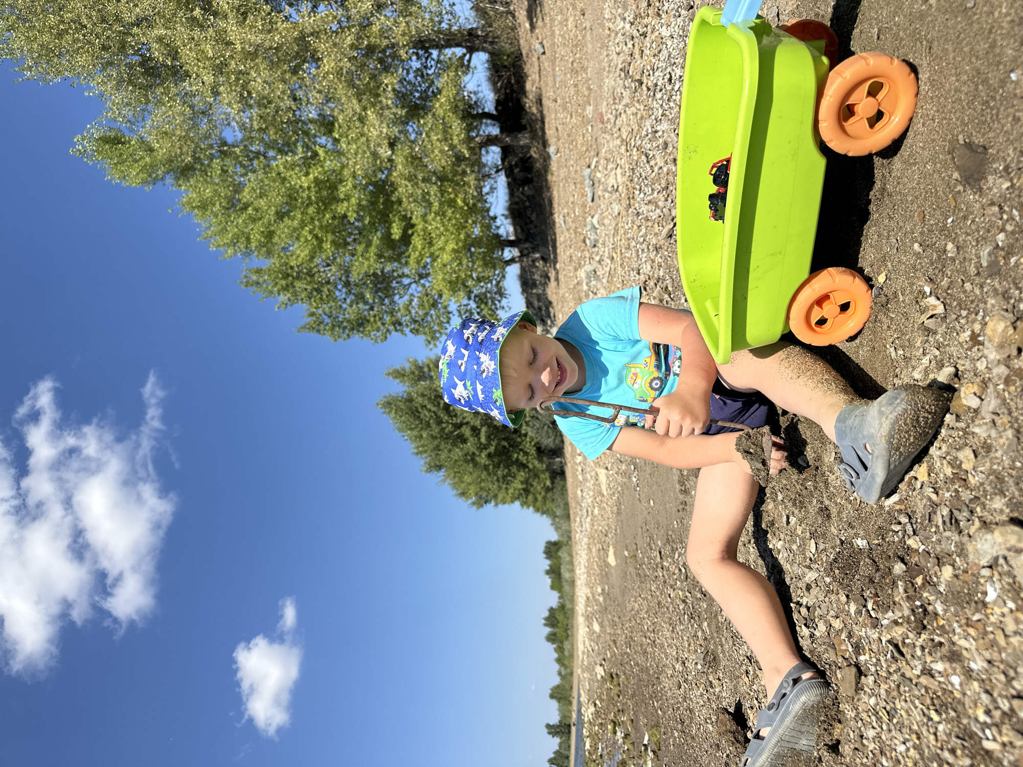 In Energetik, Russia, a young boy, wearing a blue bucket hat with a white dinosaur pattern and a teal t-shirt with a truck graphic, is sitting on a pebble-strewn beach. He holds a stick in his hands, with sand clinging to his legs and feet, which are encased in grey sandals. Beside him is a bright green toy wagon with orange wheels, containing two small toy cars. The background features a clear blue sky with scattered white clouds, and a line of trees bordering the beach. The water of a lake or river is visible on the left.
