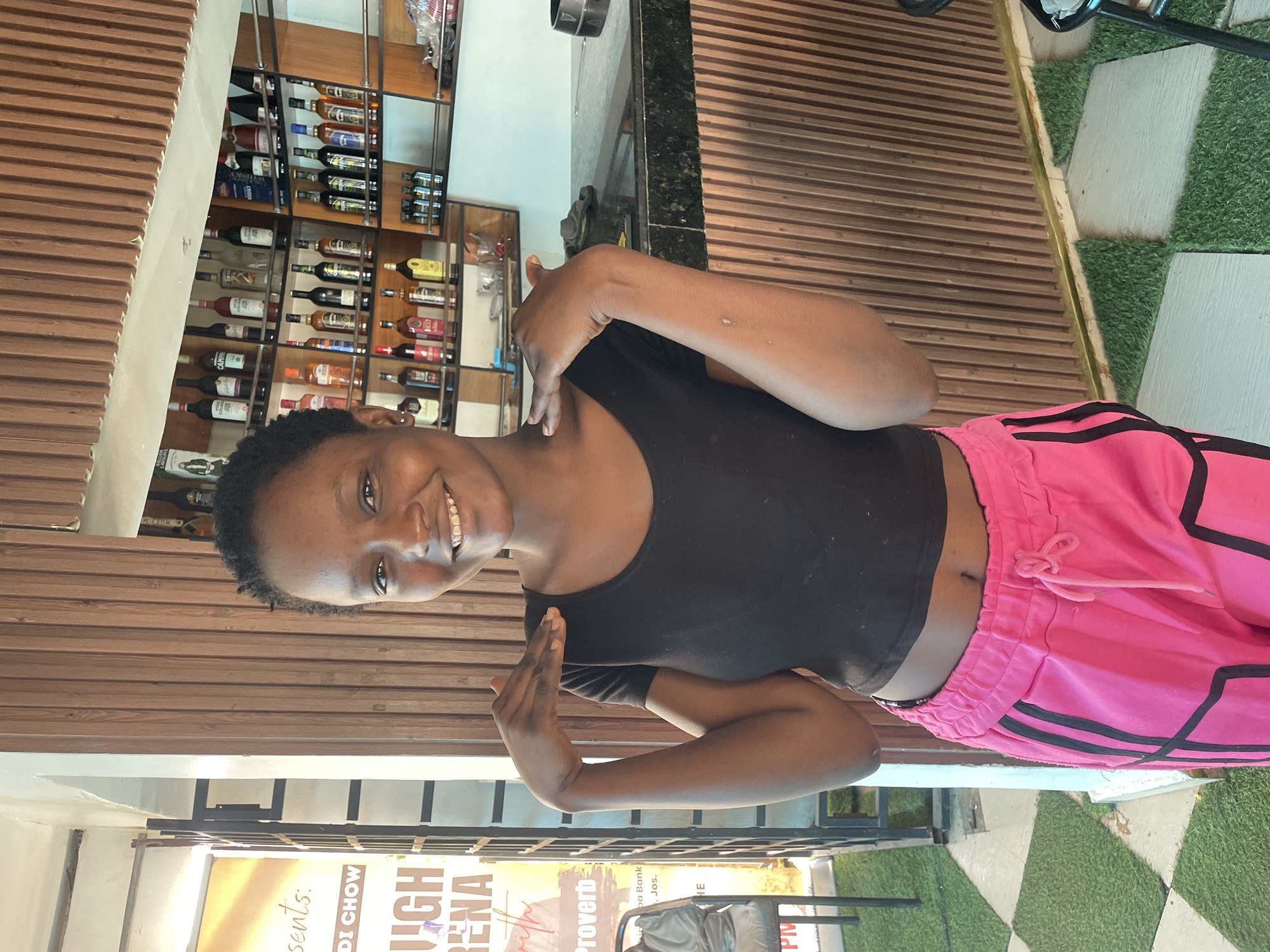 A young Black woman with short hair and a black crop top is standing in what appears to be a bar or restaurant in Jos, Nigeria. She is smiling and has her hands raised slightly, touching her shoulders. She is wearing bright pink sweatpants with black stripes. Behind her, a shelf is stocked with various bottles of alcohol. The flooring is a mix of checkered tiles and artificial grass. A sign with text is visible to the left of the woman.