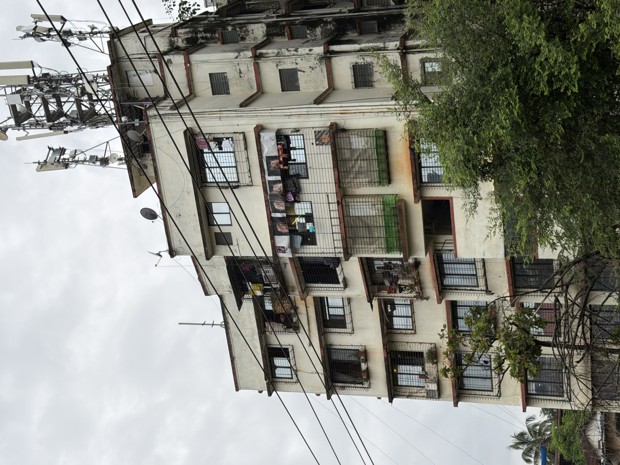 This is a full-shot, eye-level, medium-angle photograph of a weathered, multi-story apartment building in Vasai-Virar, India, on an overcast day. The building, painted in a pale, off-white color with red trim around the windows and balconies, exhibits signs of age and disrepair, with visible stains and discoloration on its facade, particularly on the right side. Numerous black wires crisscross in front of the building, obscuring parts of its exterior.
The building has many windows, most of which are covered by black metal security bars. Laundry hangs from clotheslines on several balconies. On the rooftop, a cluster of large telecommunication antennas and satellite dishes are prominently displayed. Surrounding the base of the building and extending into the foreground is lush green foliage, including trees and palm fronds, indicating a tropical or subtropical environment. The sky is uniformly gray with clouds. No people are visible in the image.
