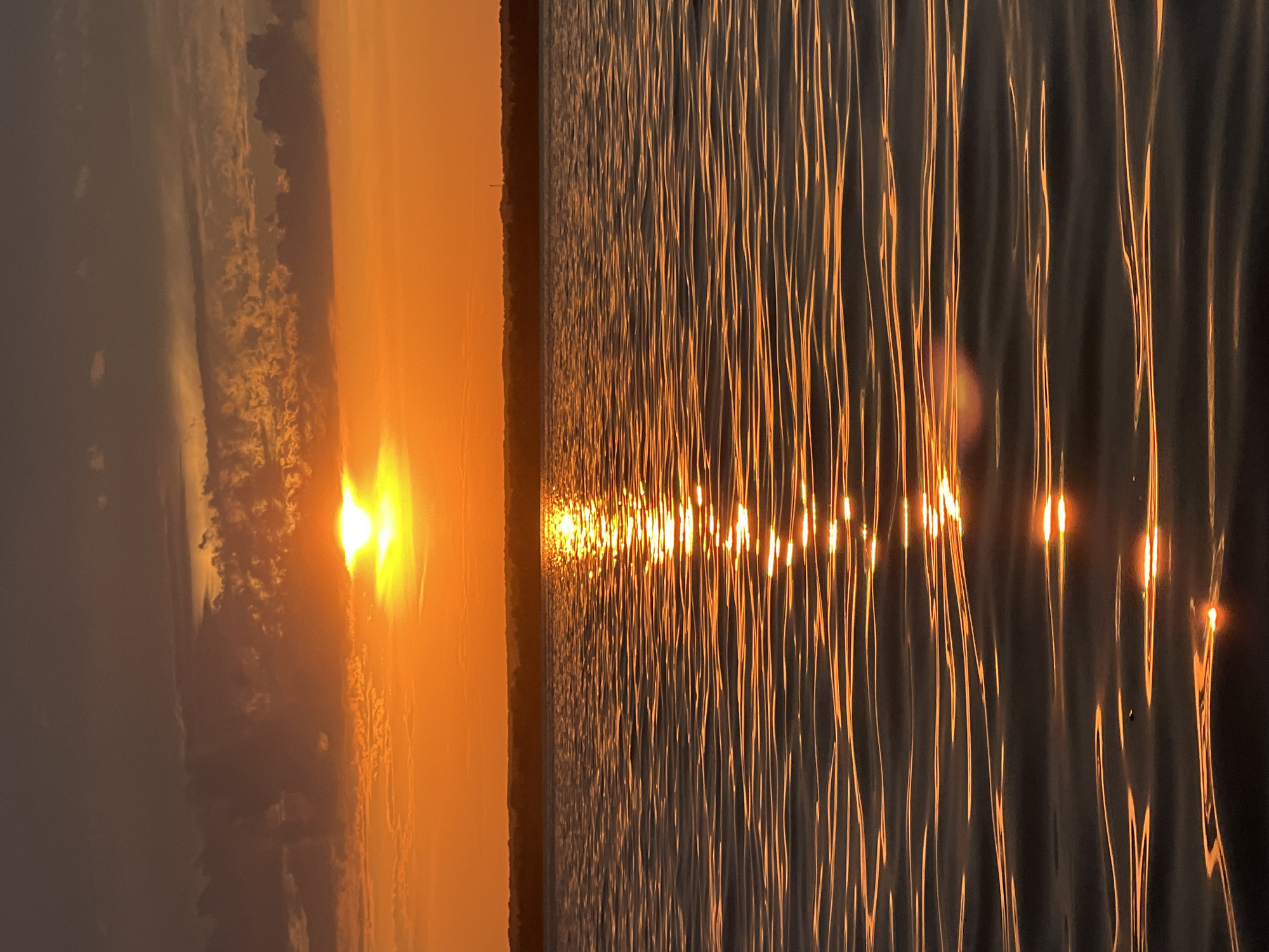 A vertical, medium shot captures a tranquil sunset over a body of water, likely a lake or sea, with the horizon line visible in the background. The sky is a vibrant gradient of orange and yellow, with the sun, a bright white orb, partially obscured by wispy, dark gray clouds in the upper left quadrant. The water's surface is rippled, reflecting the warm hues of the sunset in a shimmering path that stretches towards the viewer. The distant shoreline is a dark silhouette against the colorful sky, suggesting a wooded or hilly landscape. The image is devoid of any human presence or man-made structures, focusing solely on the natural beauty of the sunset and its reflection on the water. The atmosphere is serene and peaceful, with the warm light casting a soft glow over the entire scene. The location is identified as Beregovoe, Russia.