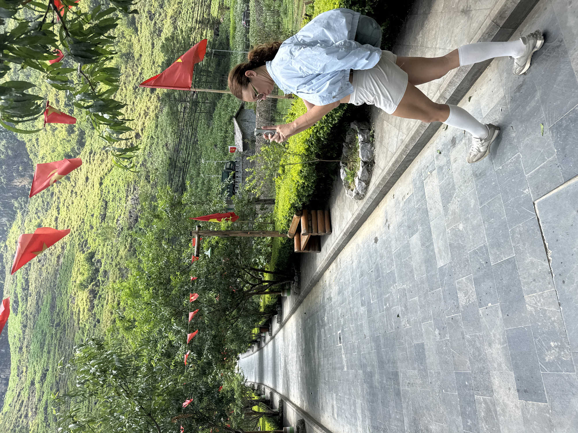 A woman in light blue jacket, white shorts and white knee-high socks stands on a stone-paved pathway, holding a phone. The pathway is lined with trees adorned with small red flags. A steep, green hillside rises to the left of the pathway. The scene is located in Mèo Vạc Commune, Vietnam.