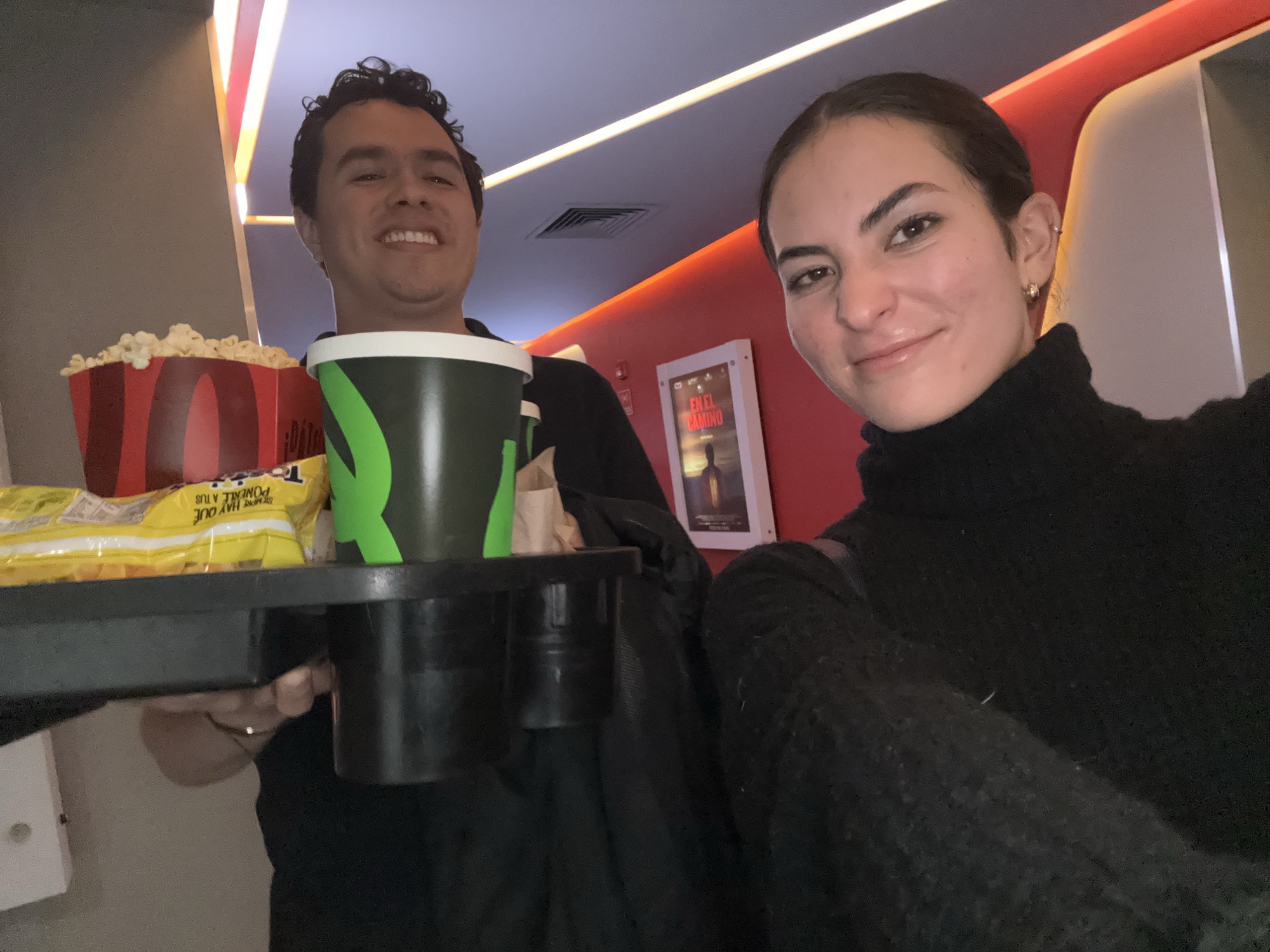 In a selfie taken in what appears to be the lobby of a movie theater, a smiling young woman with dark hair pulled back, wearing a black turtleneck sweater, holds the camera. To her left, a smiling young man with curly dark hair holds a black tray containing a large container of popcorn, a yellow bag of snacks, and a large green and black drink cup. In the background, there are red walls and lighting features. A framed movie poster is visible on the wall behind them. The theater is located in the city of Puebla, Mexico.