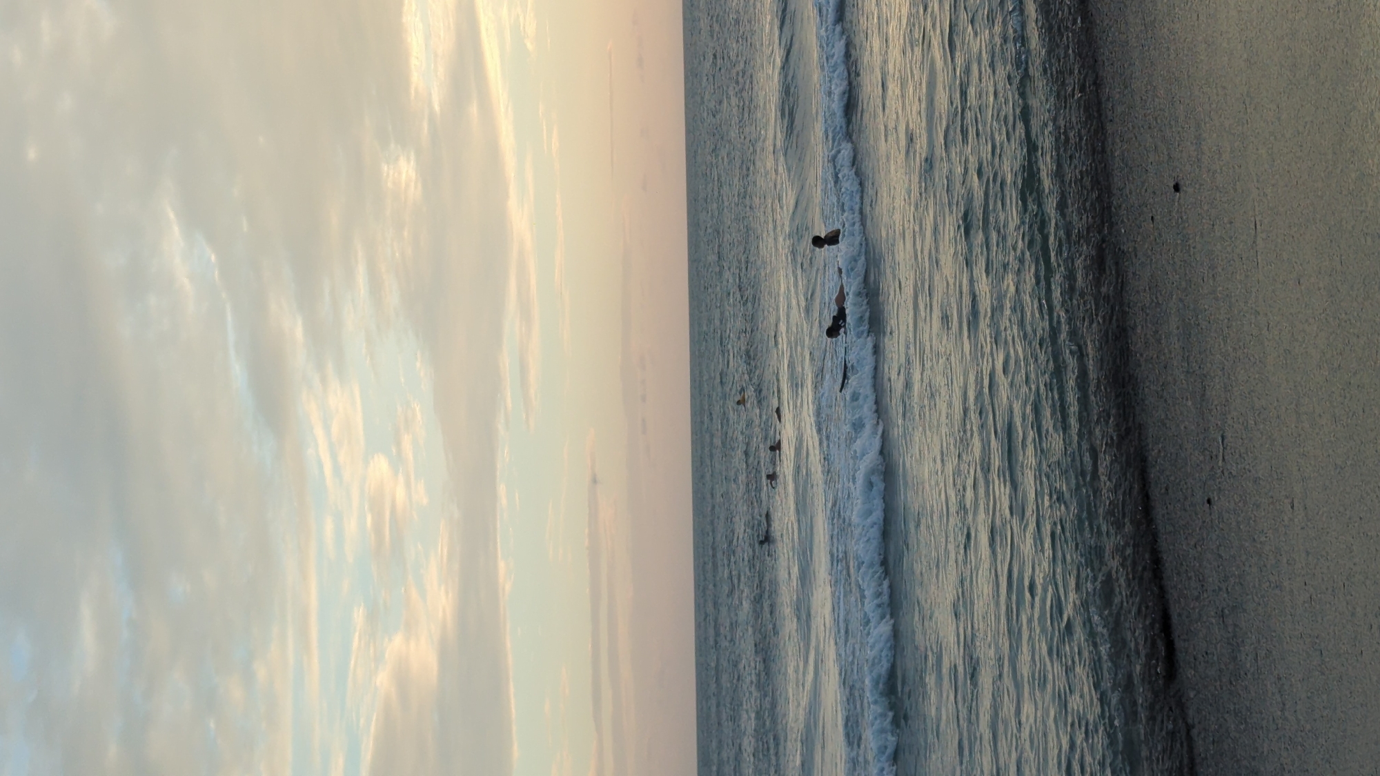 This vertical outdoor shot depicts a beach scene in North Kuta, Indonesia. The sky, occupying the left side, features white clouds tinged with gold, suggesting either sunrise or sunset. The right side of the frame showcases the ocean meeting the sandy beach. Several individuals are present in the water; two are notably positioned further out, appearing to be on surfboards, while several other smaller, silhouetted figures are closer to the shore. Waves are visible, with the foamy edges contrasting against the darker water. The sand on the right is wet and smooth, reflecting the light. A couple of darker spots are visible in the sand, potentially debris or marks.