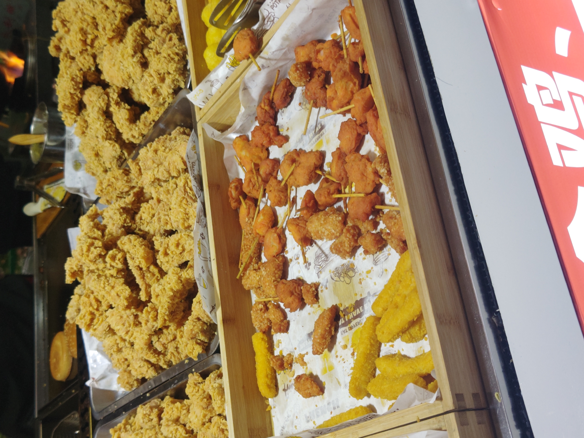 A high-angle shot displays a street food stand in Shapingba District, China. The stand presents various fried foods in trays and a wooden box. The trays contain piles of golden-brown fried chicken. The wooden box contains orange-colored fried chicken skewers, and deep-fried yellow sticks. The trays and the box are lined with white paper. Part of a red and white sign is visible to the right of the wooden box. A silver pot is visible on the left. The lighting is likely from streetlights or other artificial sources.