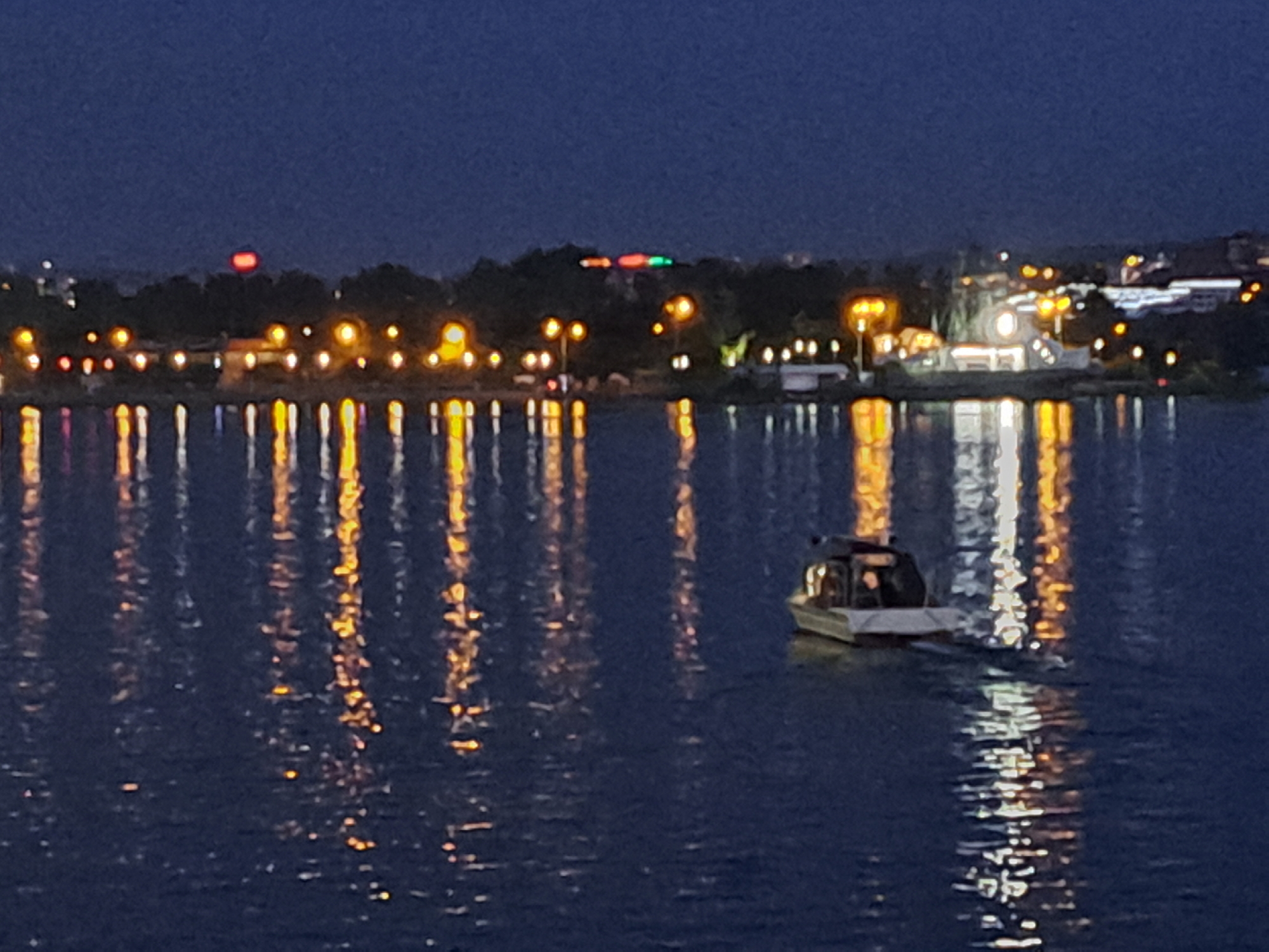 The image shows a body of water, likely a river or lake, at dusk or night. The water reflects numerous yellow and white lights from a city skyline in the background. Trees are visible along the shoreline, silhouetted against the lights. A small boat with a dark cabin is moving across the water in the foreground, leaving a wake. The sky is a deep blue. The lights on the distant shore suggest an urban environment with buildings and possibly streetlights. Without further information, it is impossible to definitively identify the specific location within Irkutsk, Russia. However, the presence of a body of water and city lights is characteristic of many waterfront areas in cities.