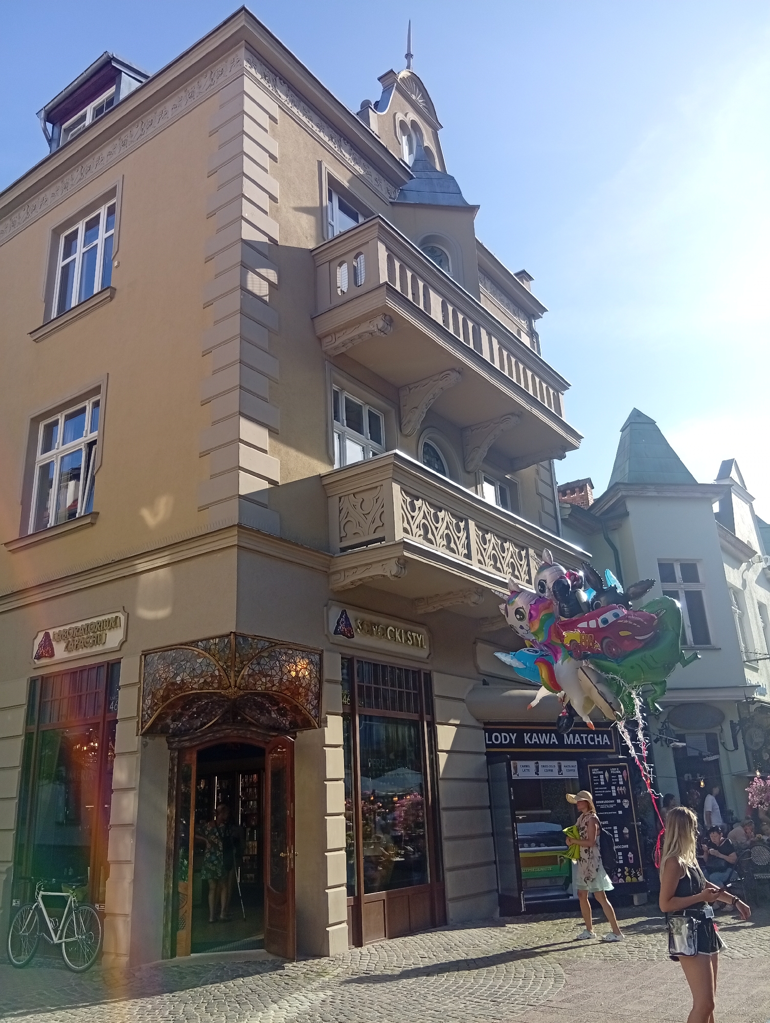 A sunny day is captured in a street scene in Sopot, Poland, featuring the corner of an ornate, tan-colored building. The building exhibits classical architectural elements, including detailed cornices, balconies with decorative railings, and large windows. The ground level of the building houses several shops, one with a decorative stained-glass awning over its entrance and another with a prominent sign reading "LODY KAWA MATCHA" (Ice Cream Coffee Matcha).
In the foreground, a white bicycle is parked to the left of the entrance. A cluster of colorful helium balloons, including a dinosaur, a car, and other cartoon characters, floats near the "LODY KAWA MATCHA" stand. Several people are visible: a woman wearing a hat and a floral dress stands near the ice cream stand, holding green items; another woman in a black top and shorts is further to the right, looking at her phone. In the background, other pedestrians and outdoor seating areas of businesses suggest a lively urban environment. The street is paved with cobblestones, and shadows are cast by the buildings, indicating the sun's position. The sky is clear blue.