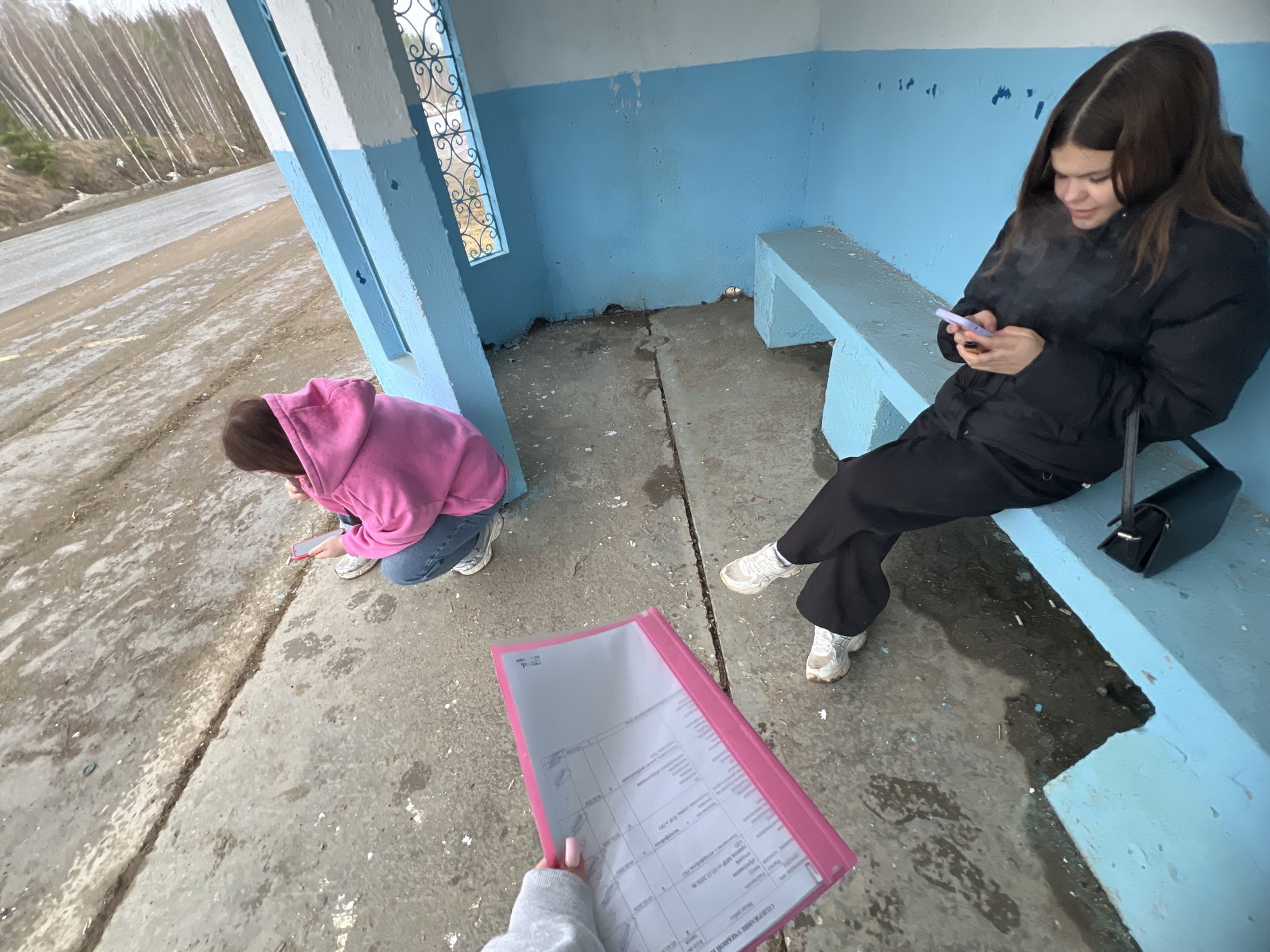 Two young women are at a bus stop in what appears to be the city of Chaikovsky, Russia. One woman, wearing a black puffer jacket and black pants, sits on the blue concrete bench at the bus stop, looking at her cell phone. A black purse rests on the bench beside her. The second woman, wearing a pink hoodie and jeans, kneels on the concrete floor, also looking at a cell phone. Another individual is holding a pink folder containing documents. The bus stop has blue painted walls and white painted columns. The surrounding area includes a road and trees.