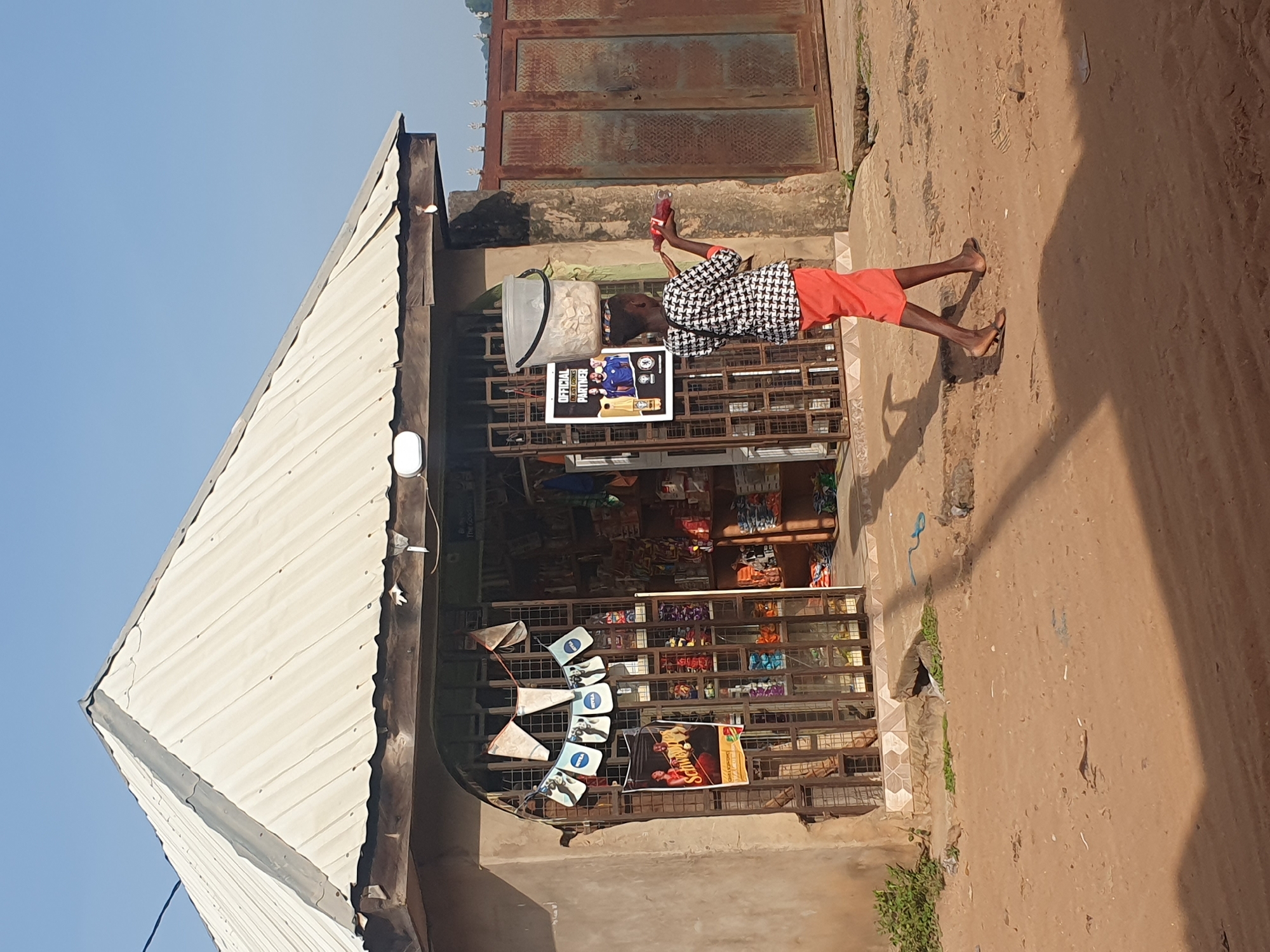 The image depicts a street scene in Lokoja, Nigeria, featuring a small storefront with goods displayed behind a metal grate. A young person, dressed in a black and white patterned shirt and an orange skirt, walks away from the store, carrying a large clear plastic bucket filled with white items on their head. They are also holding a red bottle in their right hand. The storefront has a corrugated metal roof and a light-colored facade. A banner with "OFFICIAL PARTNER" is visible inside the store. The ground is dusty, characteristic of a dry environment. The background includes a rusty metal gate and a clear blue sky. The scene appears to be during daylight hours.