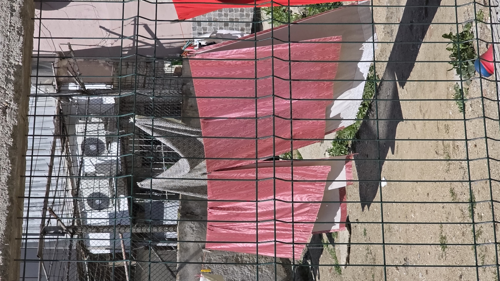 A fenced-off outdoor area, likely in Blagoevgrad, Bulgaria, based on the building style and vegetation. A wire fence is in the foreground. Behind it, laundry is hung out to dry, including red and white fabric, and a patterned gray and white textile. A building with multiple air conditioning units is visible in the background. The ground is dirt or gravel, with some grass. A shadow is cast across the scene. The image is taken from an elevated viewpoint, looking down on the scene.